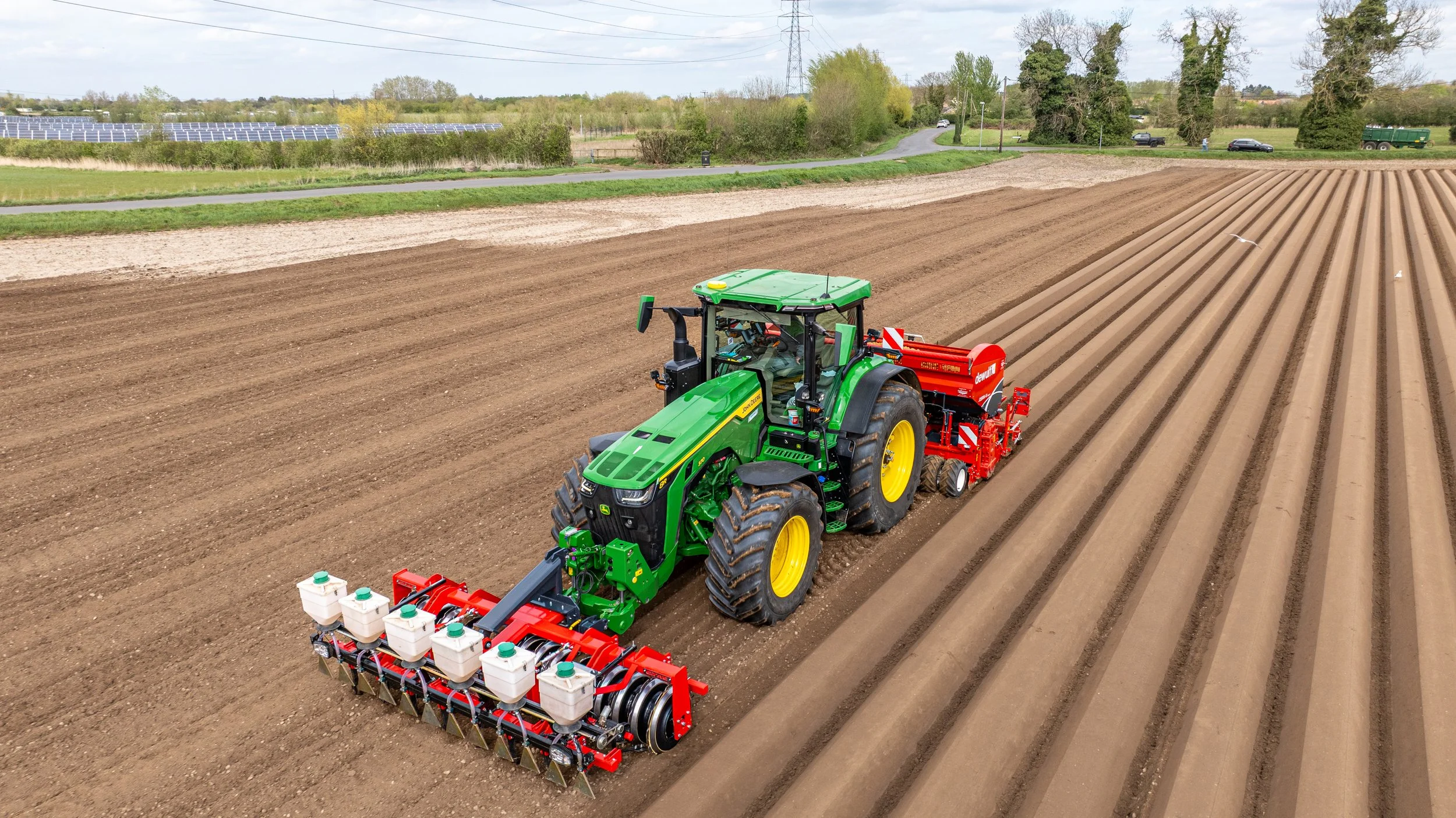 A green tractor with yellow wheels working in a field, planting seeds in freshly tilled soil, with solar panels and trees in the background.