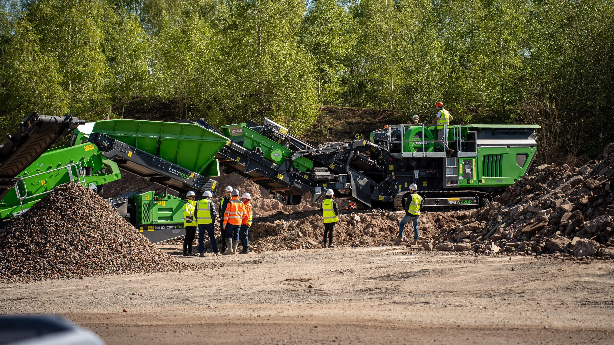 Construction workers in safety vests and helmets operating a large green rock processing machine on a gravel and rocky construction site surrounded by trees.