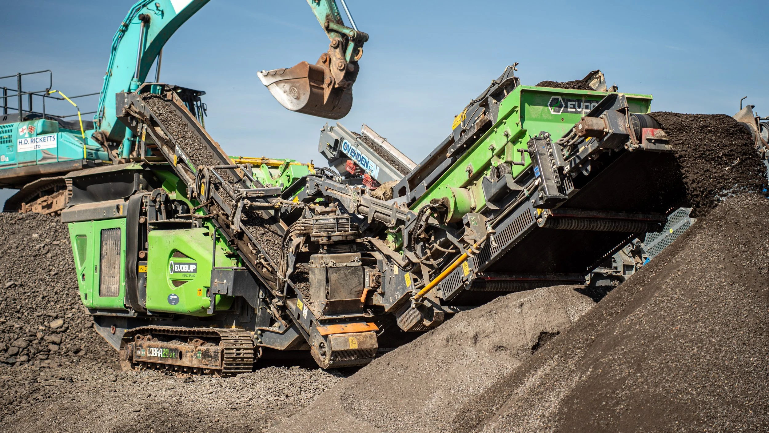 A large green and black gravel sorting machine on a dirt pile, with a blue excavator pouring gravel into it under a clear blue sky.