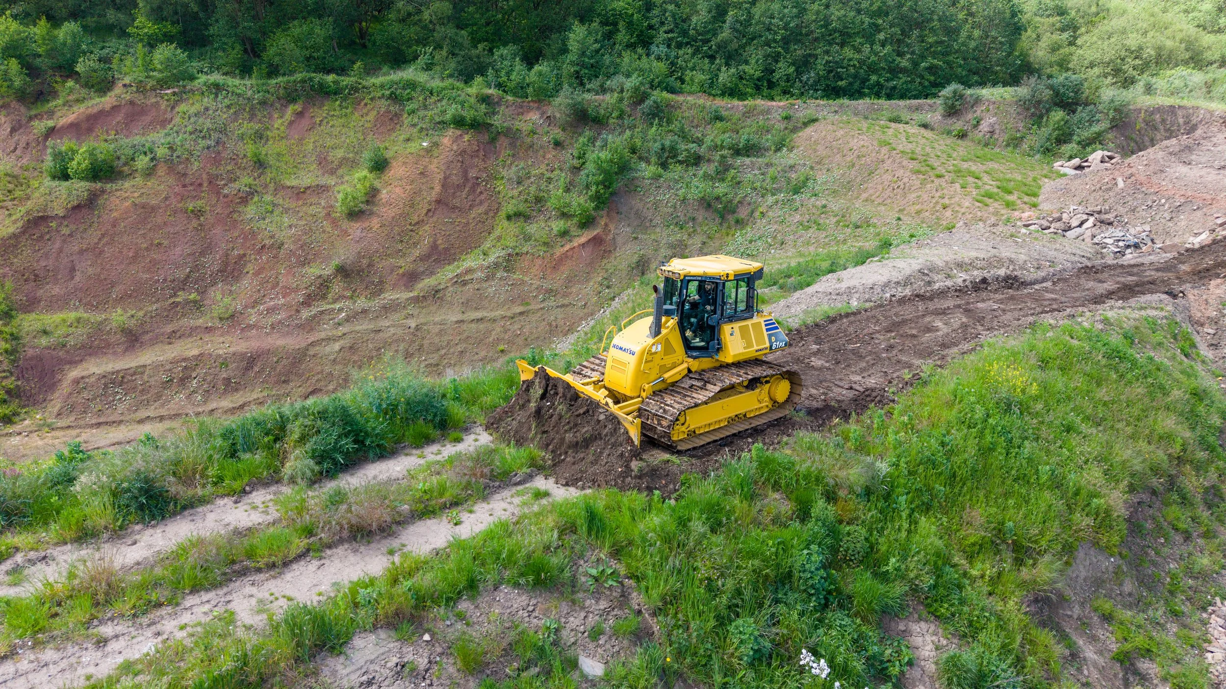 A yellow bulldozer moving dirt on a grassy hillside with trees in the background.