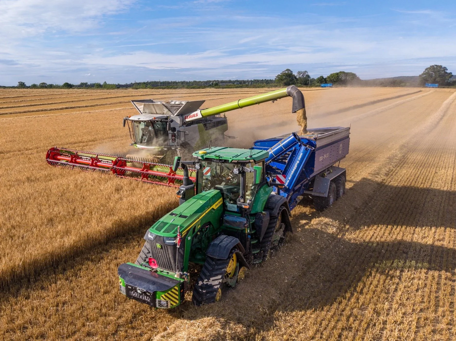 A large John Deere tractor with a red attachment harvesting wheat in a vast golden field. Two conveyor systems are transferring harvested wheat into a blue grain cart. In the background, a combine harvester harvests wheat, with blue trucks on the road.