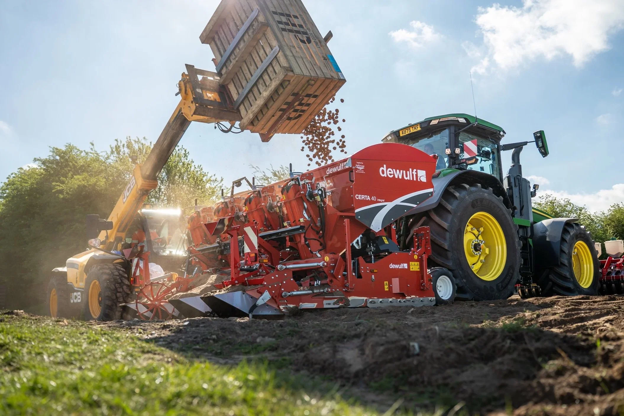 A tractor is unloading soil or gravel from a large bucket elevated in the air, with other farm machinery visible nearby and trees in the background under a partly cloudy sky.