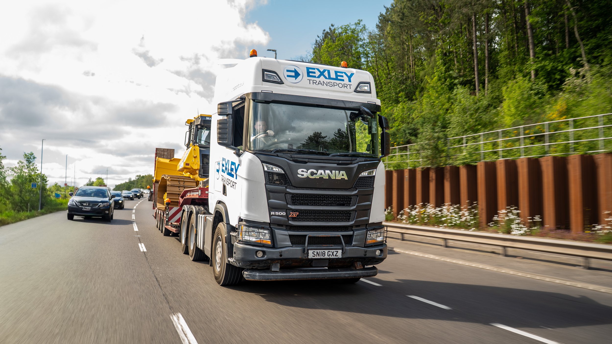A large white truck with 'Exley Transport' written on it driving on a highway on a partly cloudy day, with other vehicles visible in traffic and a green, forested hillside on the right.