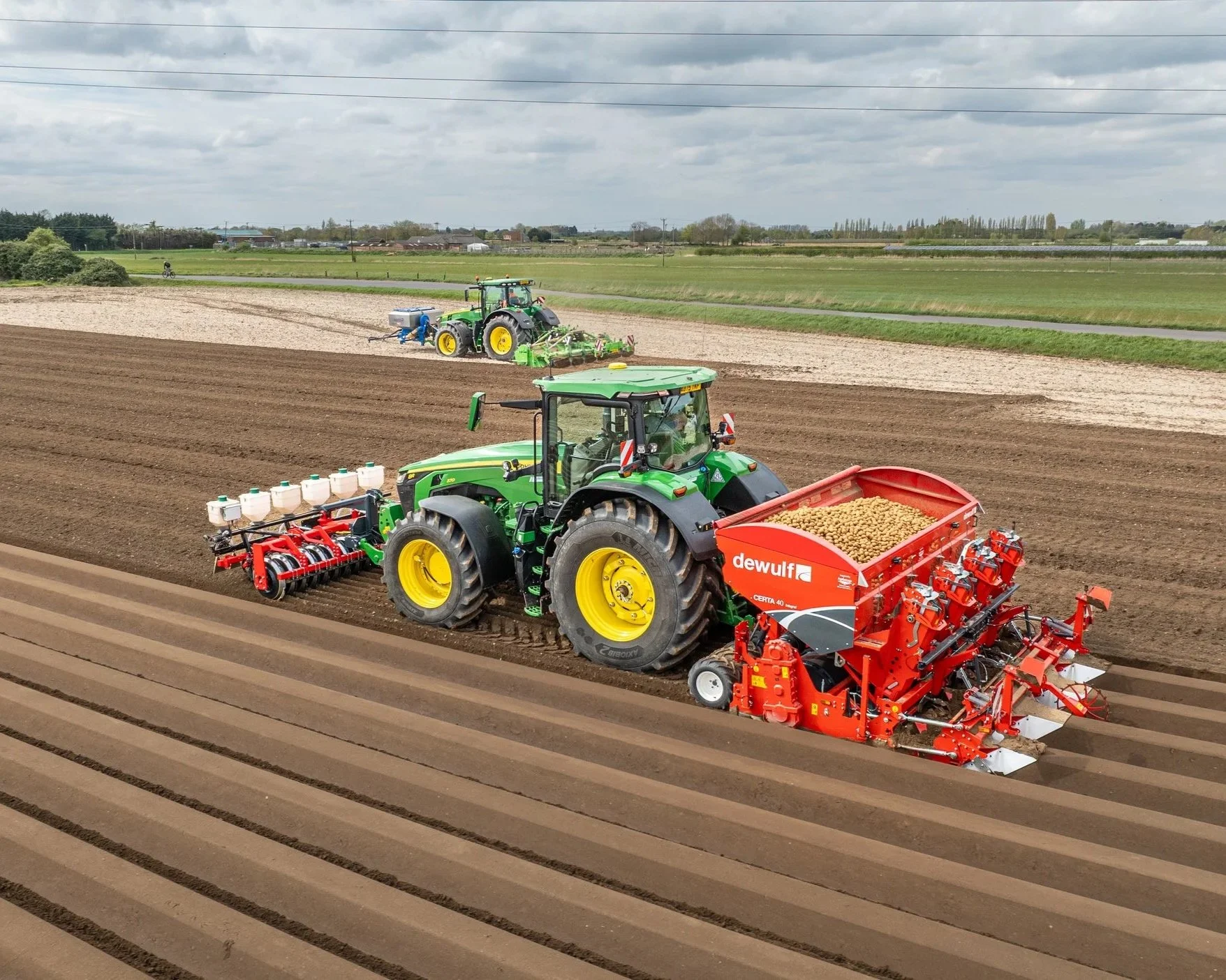 Two tractors working in a field, one planting potatoes and the other preparing the soil, with a background of green fields and a cloudy sky.