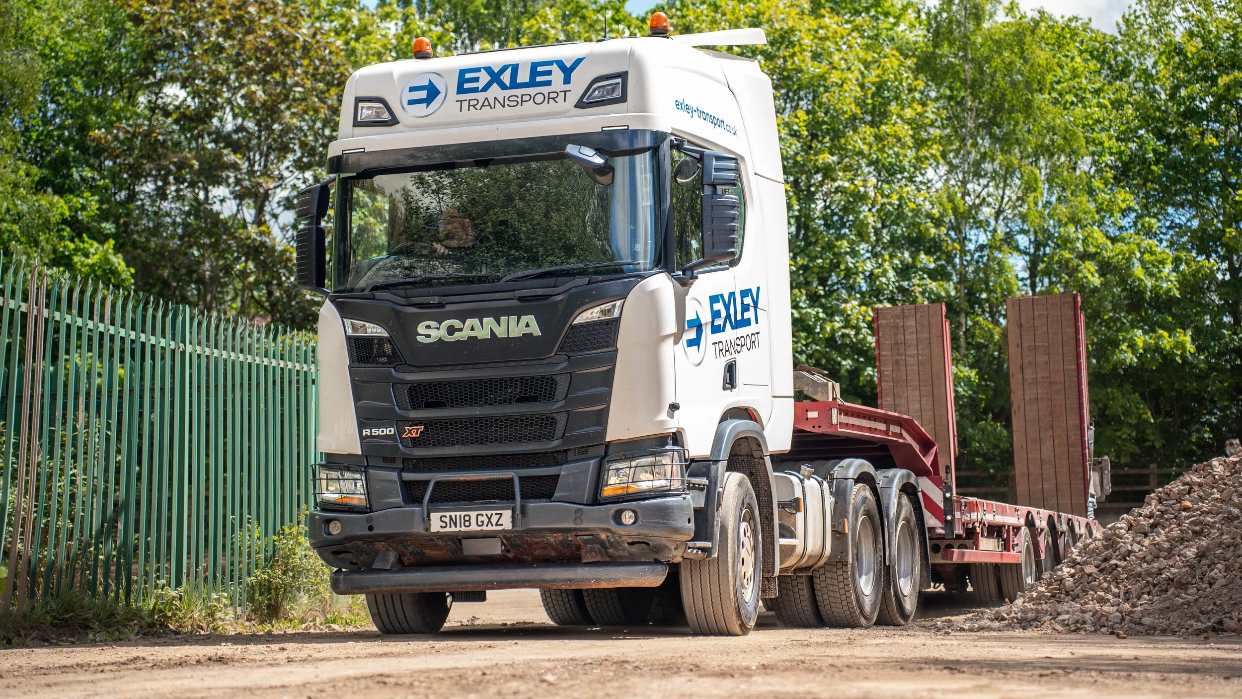 A white Scania truck with an exley transport logo on the front and sides, parked on a construction site with a pile of rocks, green trees, and a blue sky in the background.
