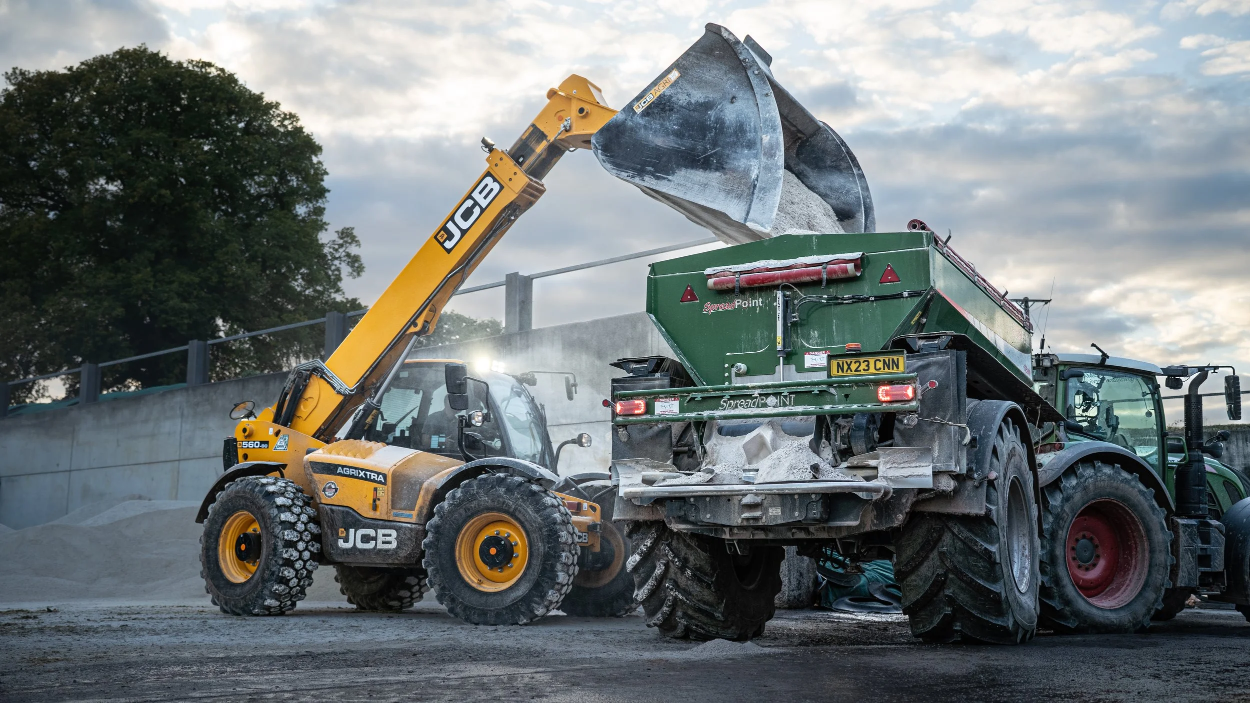 Construction site scene with a JCB excavator unloading gravel into a green spreader machine, with a cloudy sky and trees in the background.
