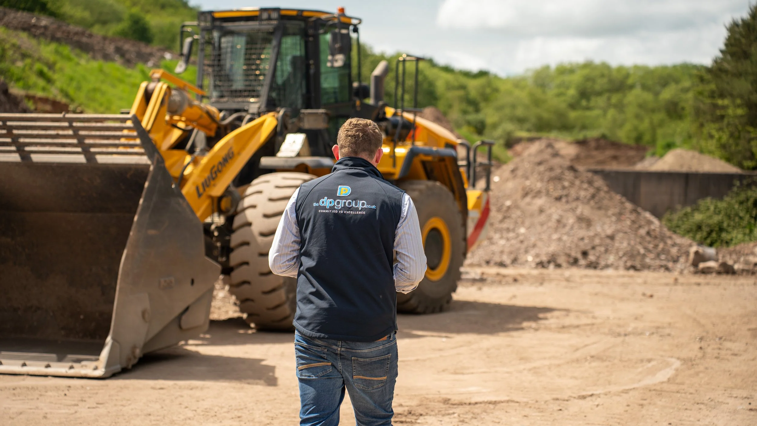A person wearing a dark vest with the logo 'dp group' on the back is walking towards a large yellow construction vehicle on an outdoor construction site with dirt and greenery in the background.