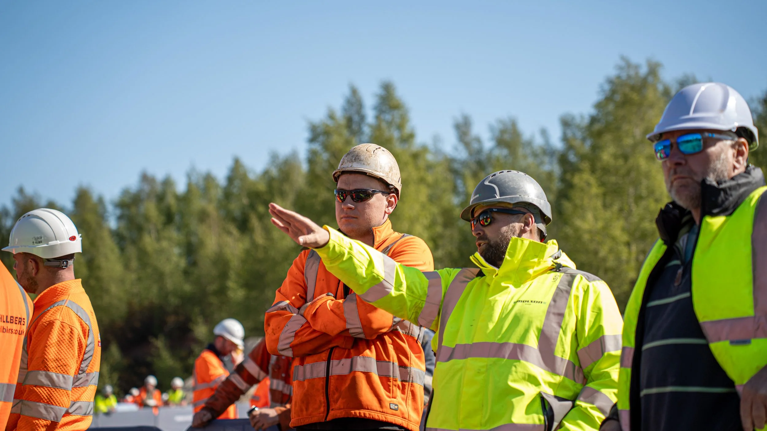 Group of construction workers wearing safety helmets and high-visibility jackets, discussing outdoors with trees in the background.