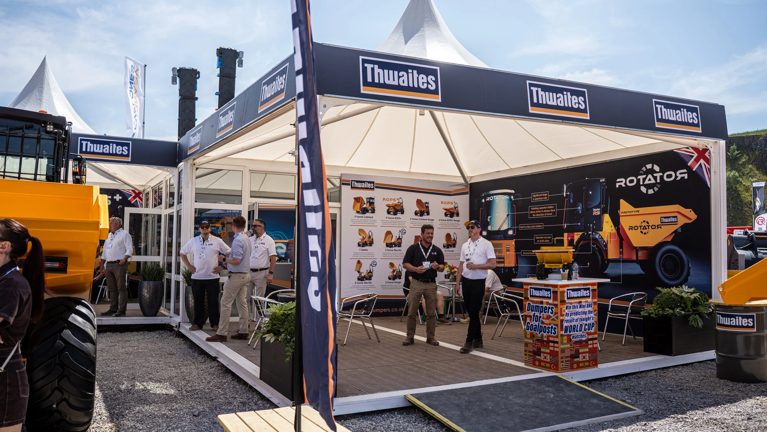 Trade show booth for Thwaites with displays of construction equipment, including a large yellow dump truck and a posters showing various machinery and specifications, with several people talking and walking around outside under a white canopy.