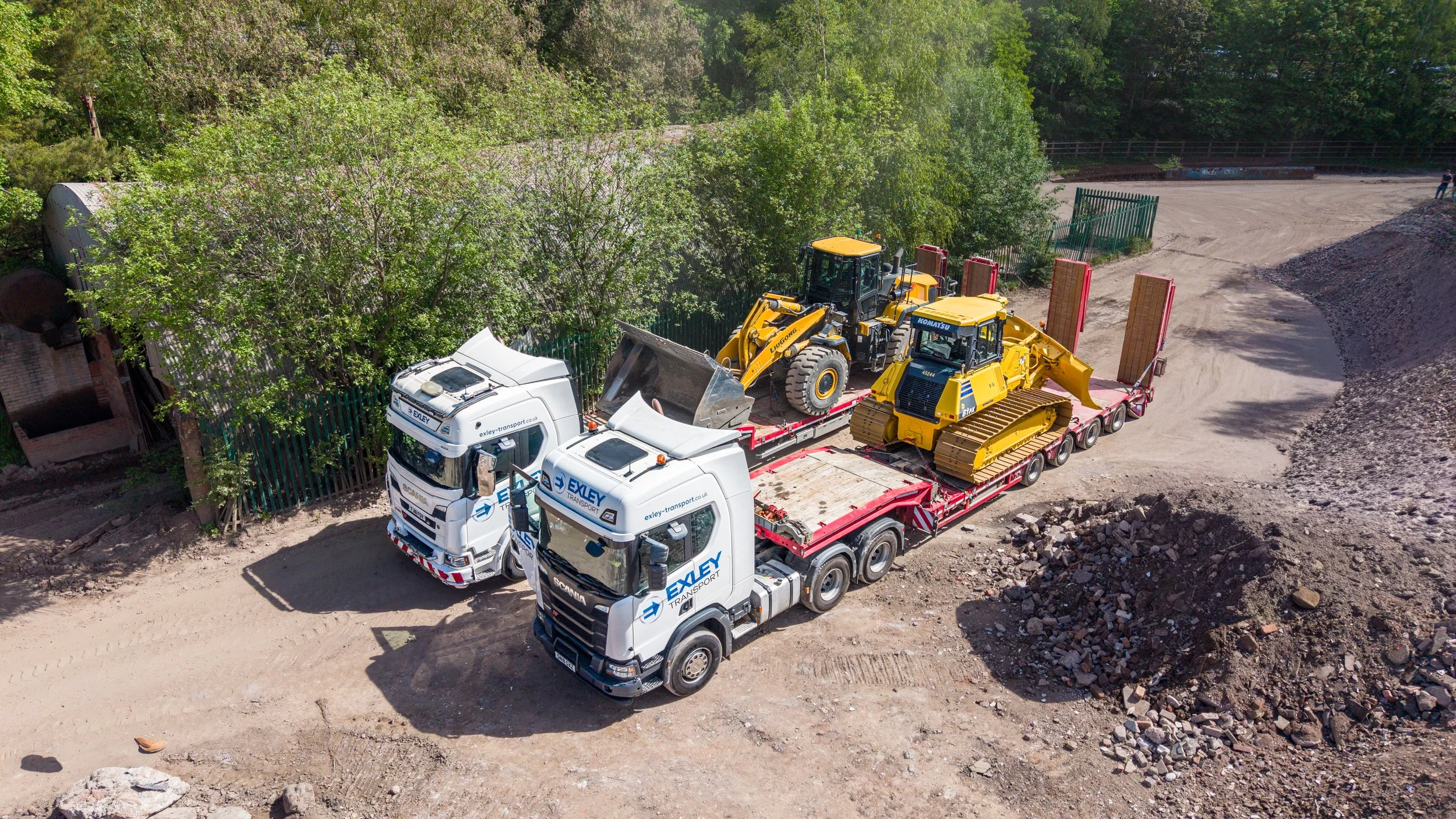 A flatbed truck transporting yellow construction machinery, including excavators, at a construction site with dirt, rocks, and green trees in the background.