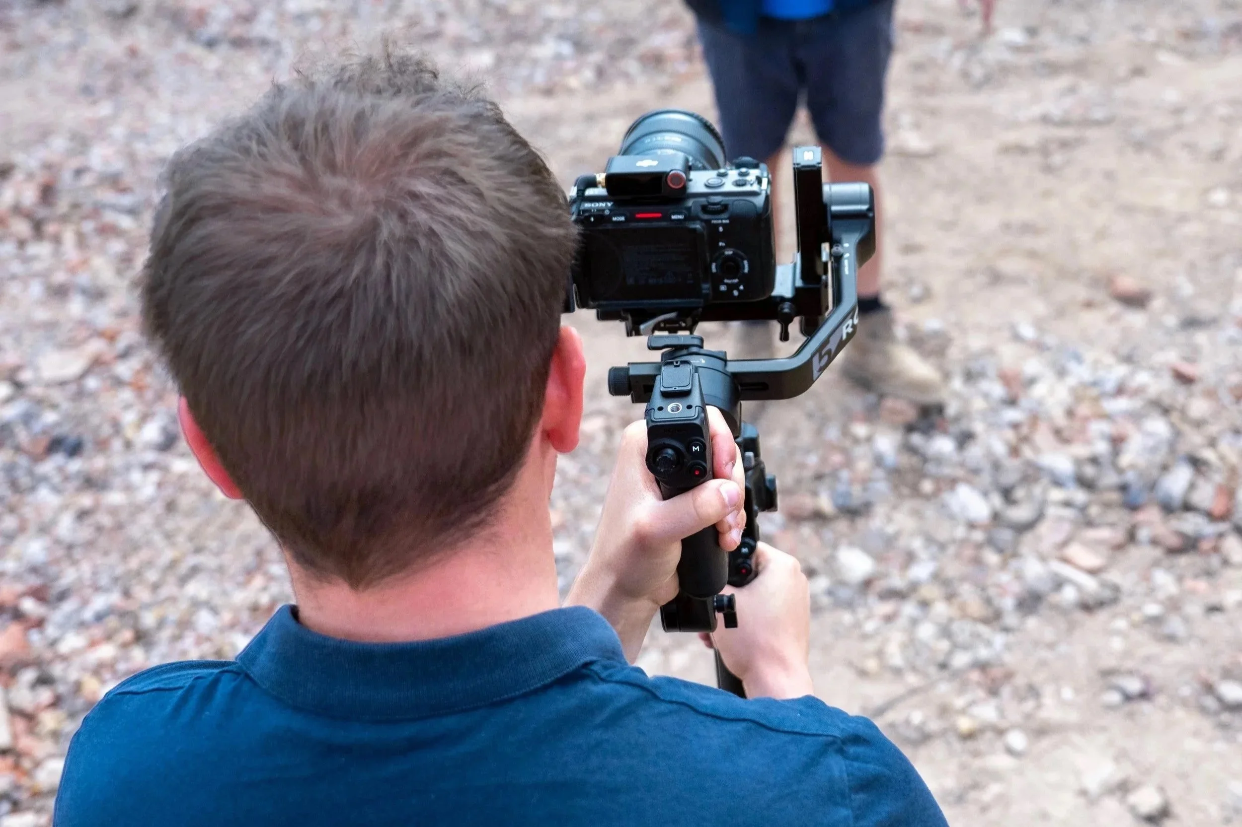 A person with short brown hair taking a photo with a camera mounted on a tripod outdoors on rocky ground.