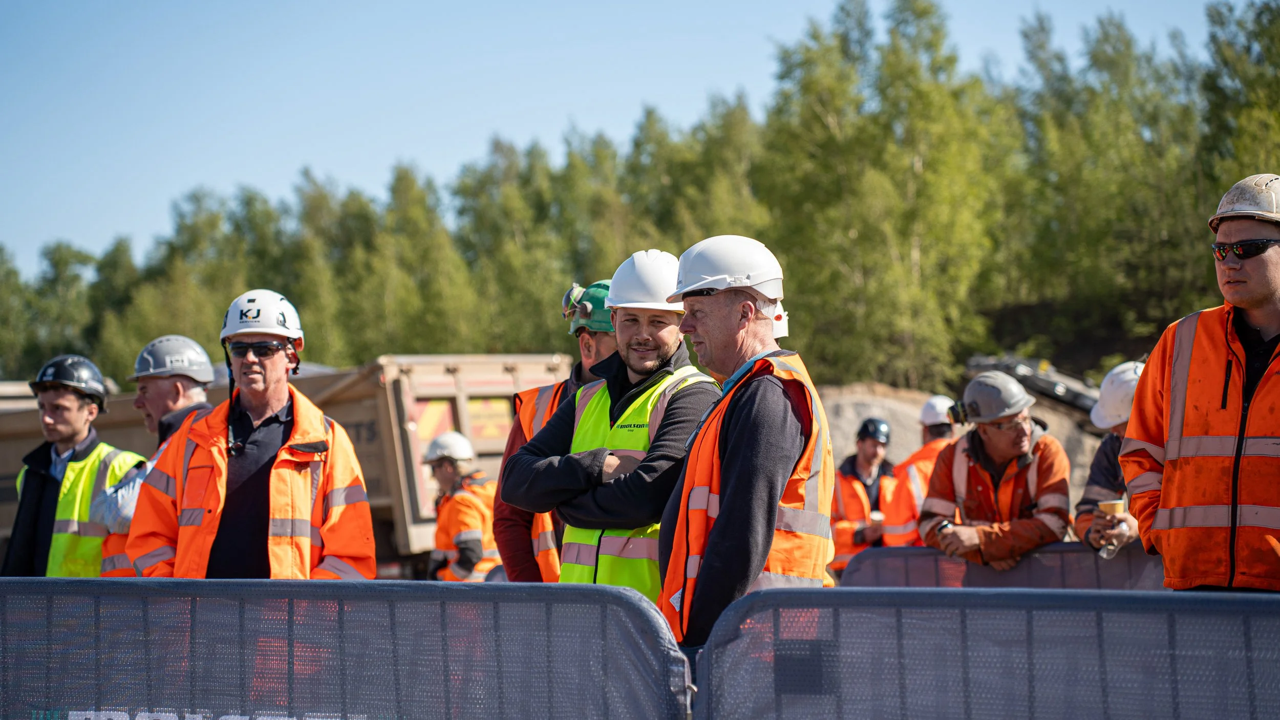 Group of construction workers wearing safety helmets and orange safety vests standing outdoors on a sunny day, with trees in the background.