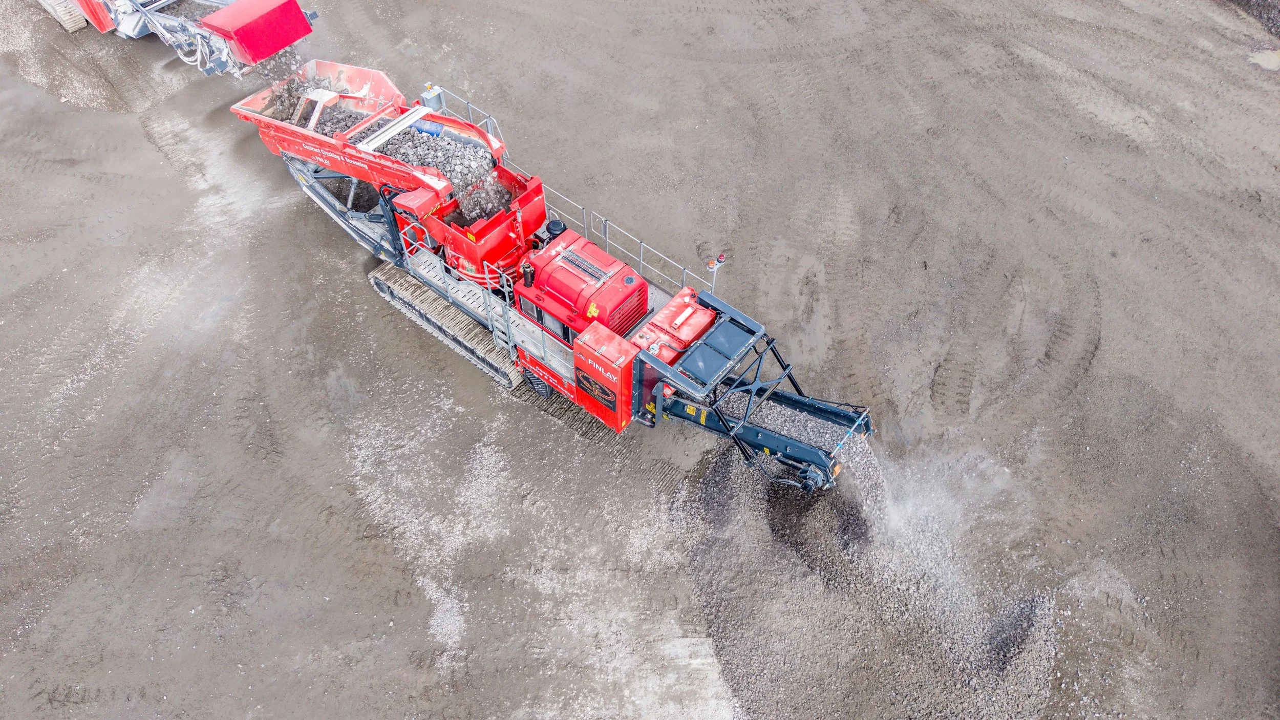 Aerial view of a large red construction machine spreading gravel on a dirt surface at a construction site.