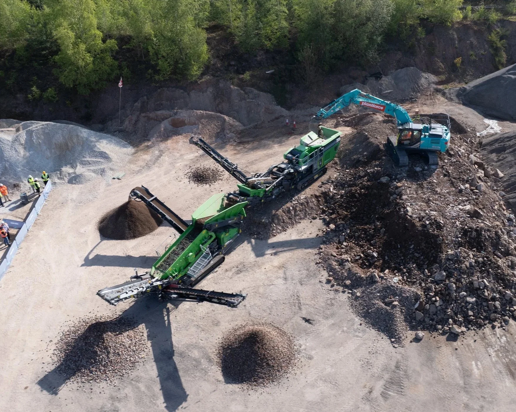 Construction site with heavy machinery, including a green conveyor belt, a large green crusher, and a blue excavator, along with workers in safety vests and helmets, surrounded by piles of rocks and dirt, and a forested area in the background.