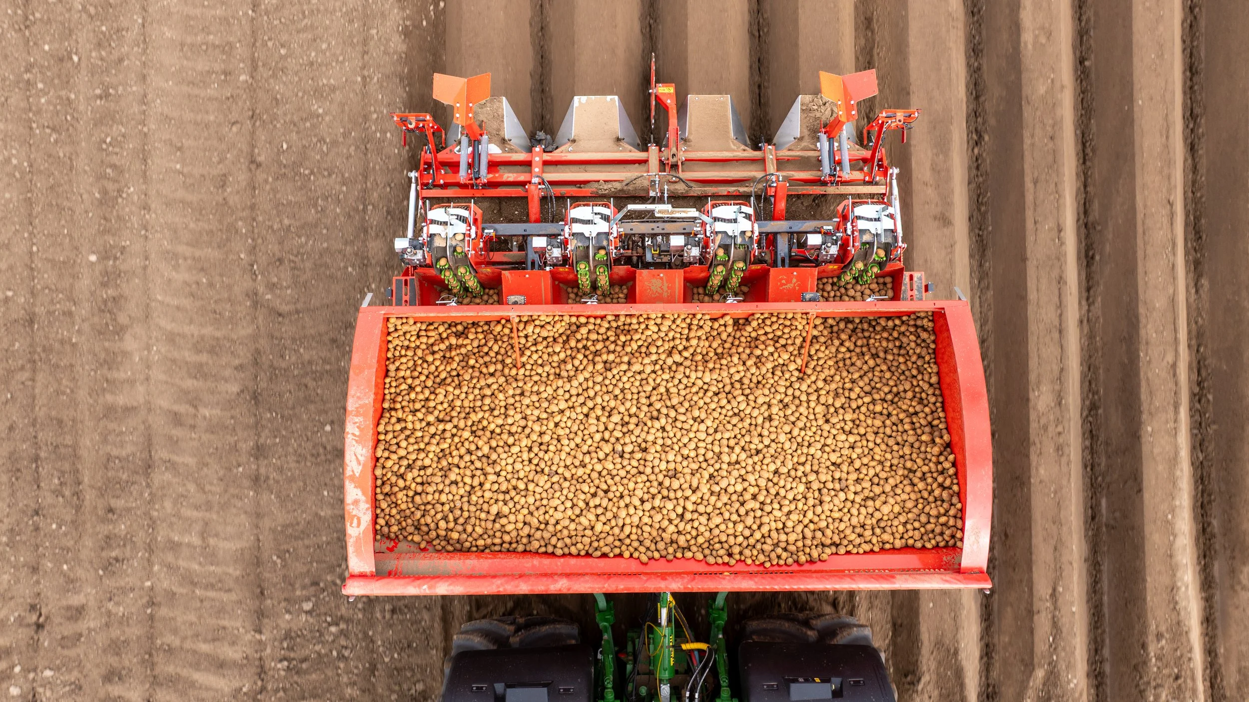 Overhead view of a red agricultural machine planting small round seeds in a field with neatly furrowed soil.