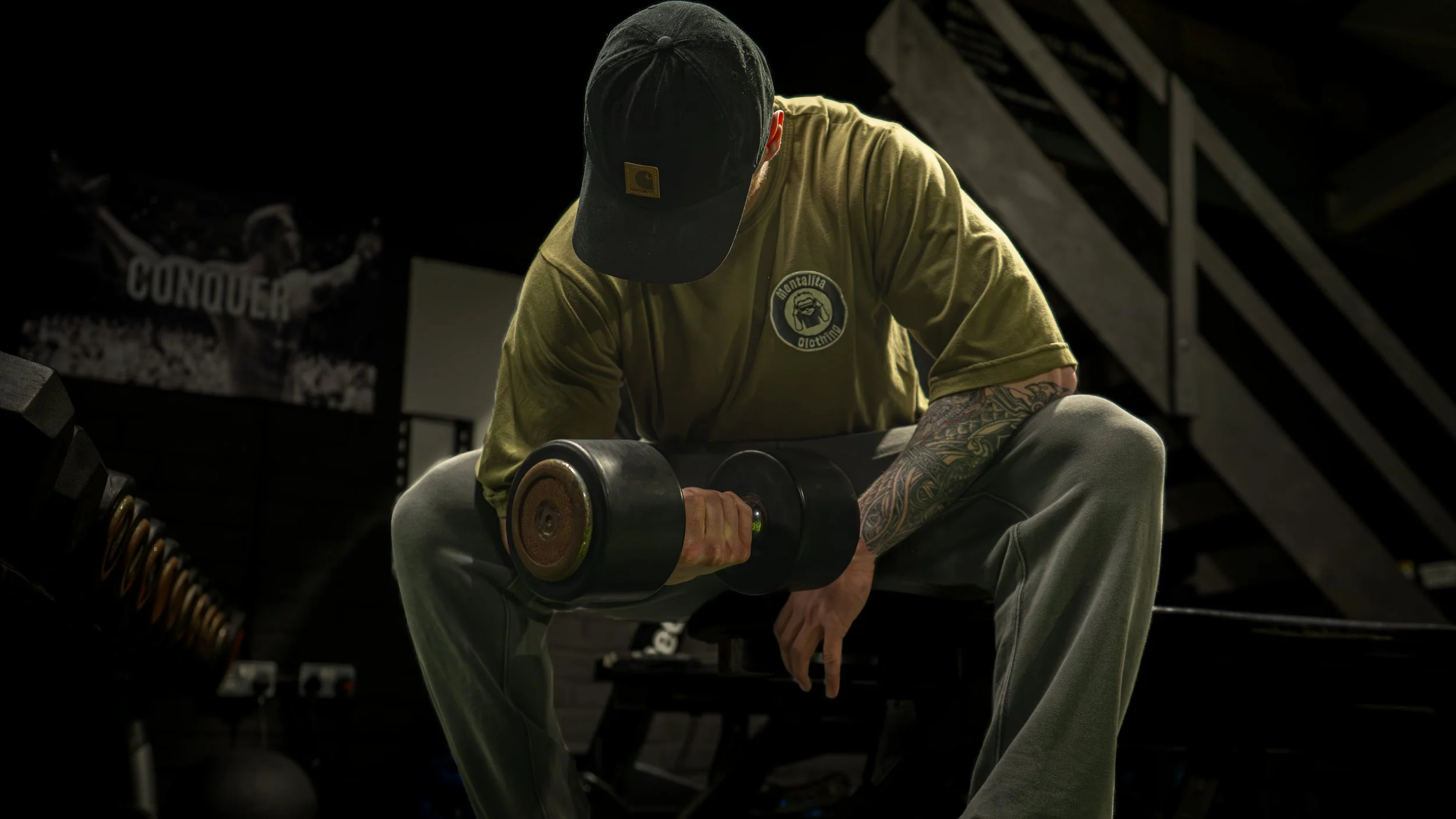 A man with tattoos wearing a black cap and khaki t-shirt, sitting on a stool lifting a dumbbell in a gym, with a dark background and a poster of a woman holding a sign that says 'Conquer'.