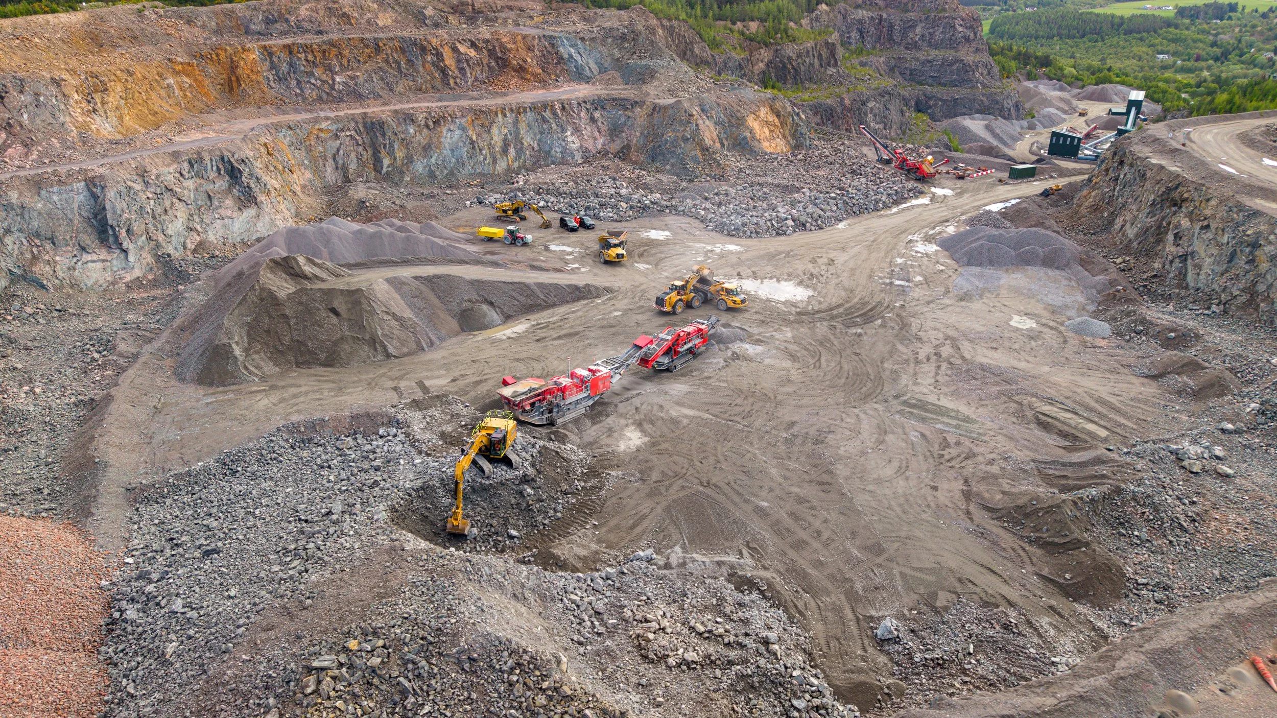 An open-pit mining site with heavy machinery, trucks, and excavators working among large piles of earth and rock, surrounded by mountainous terrain and greenery in the background.