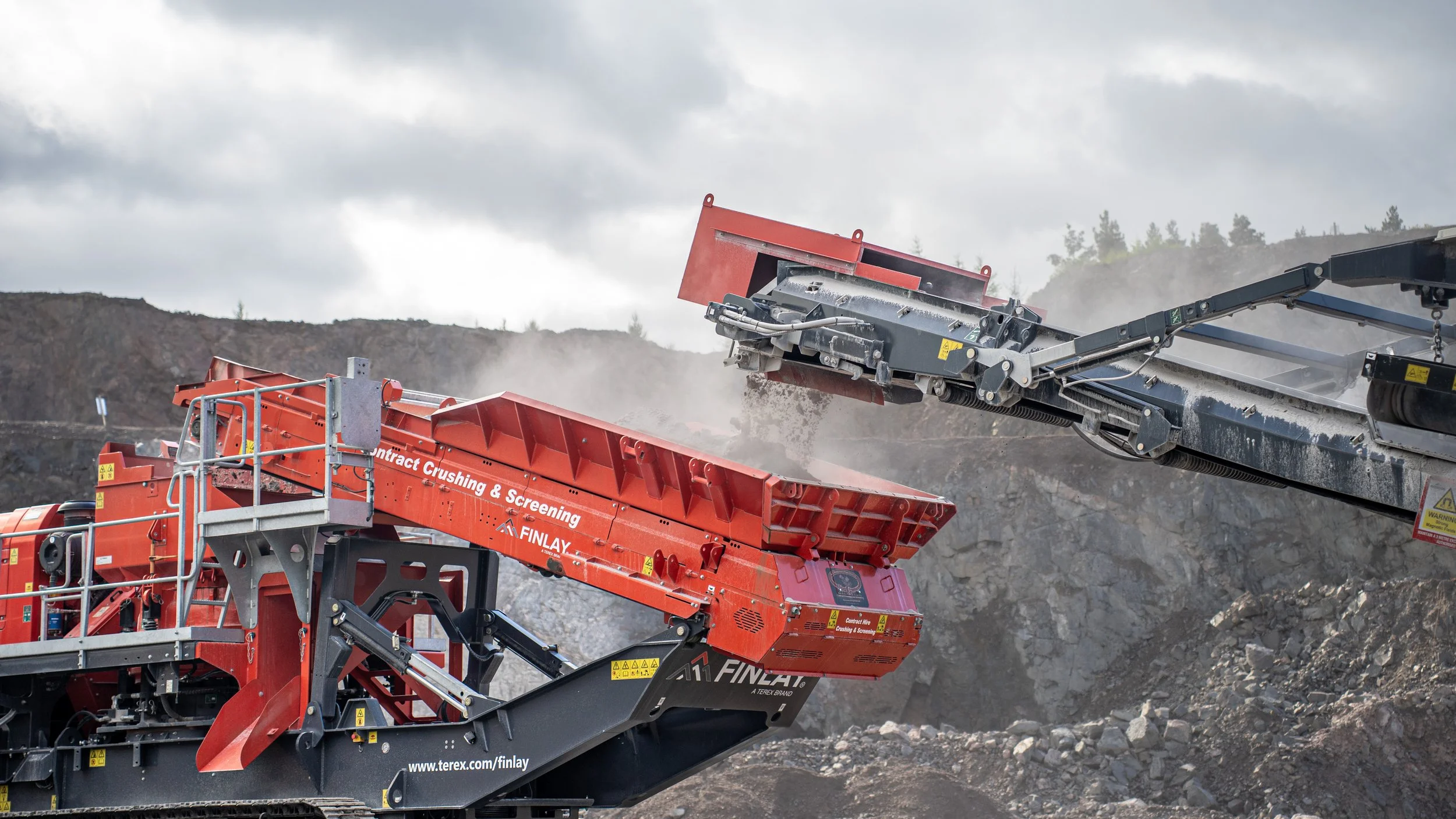 A large orange and black industrial rock crusher machine labeled 'Contract Crushing & Screening' operating outdoors on rocky terrain against a cloudy sky.