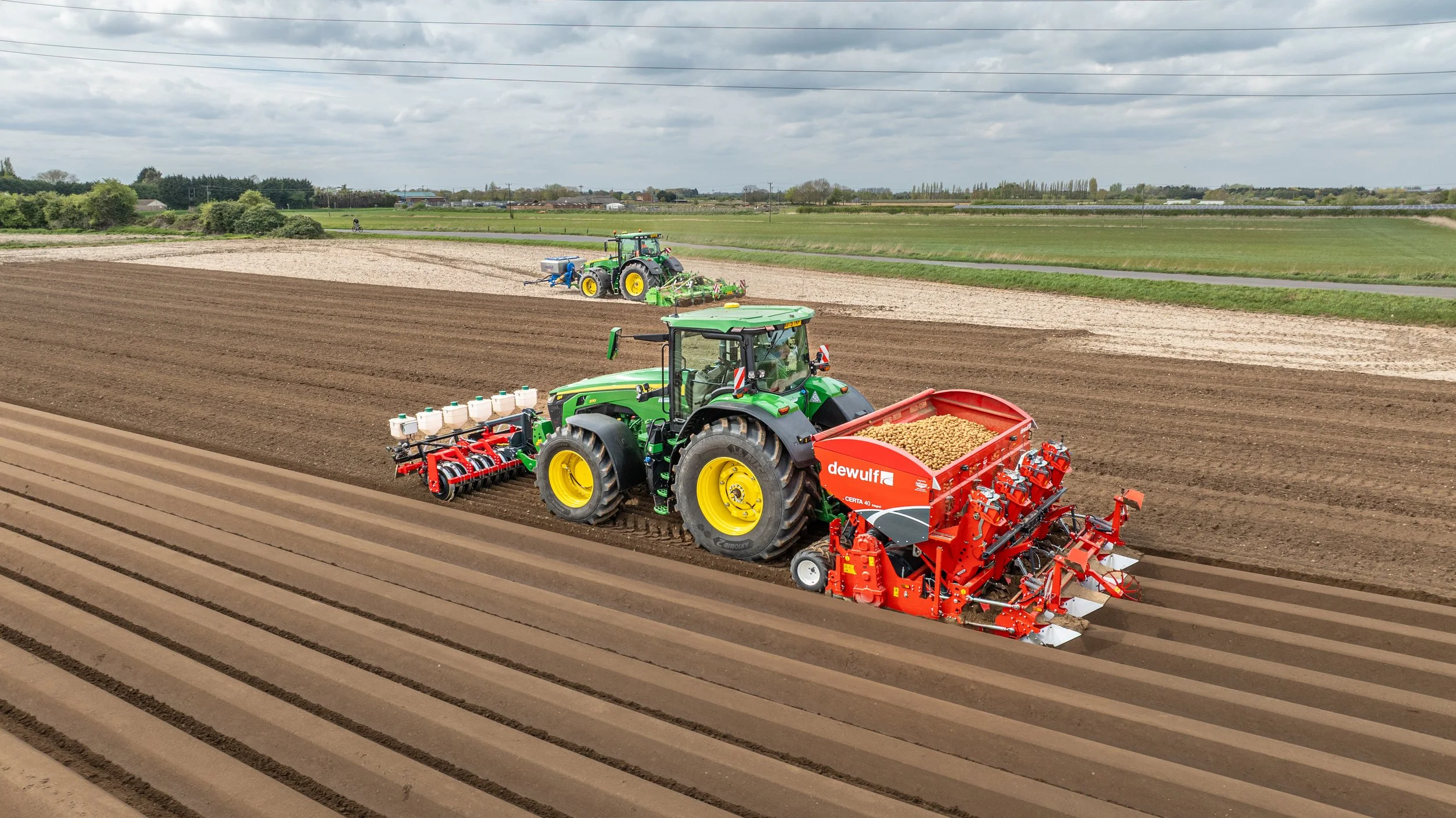 Two green tractors working in a large farm field, planting seeds or crops on freshly tilled land, with rural landscape and cloudy sky in the background.