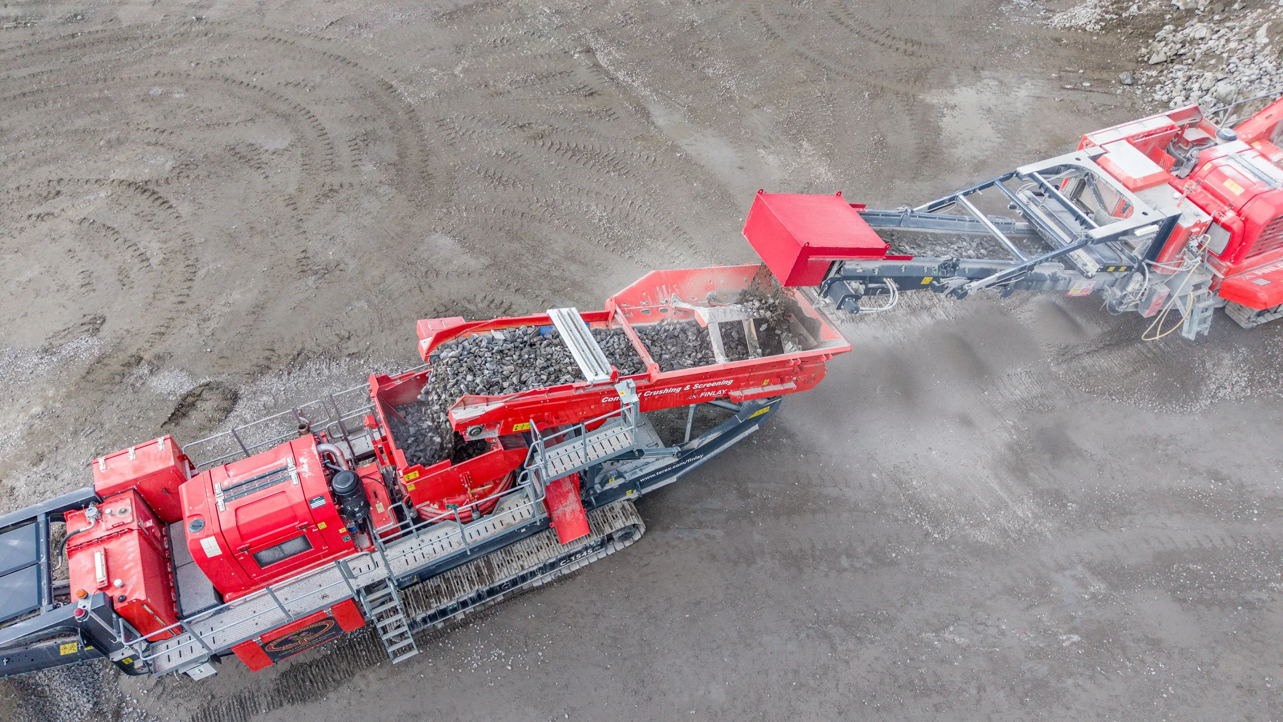 An aerial view of a red mining or construction machine with conveyor belts transporting rocks on a dirt surface.