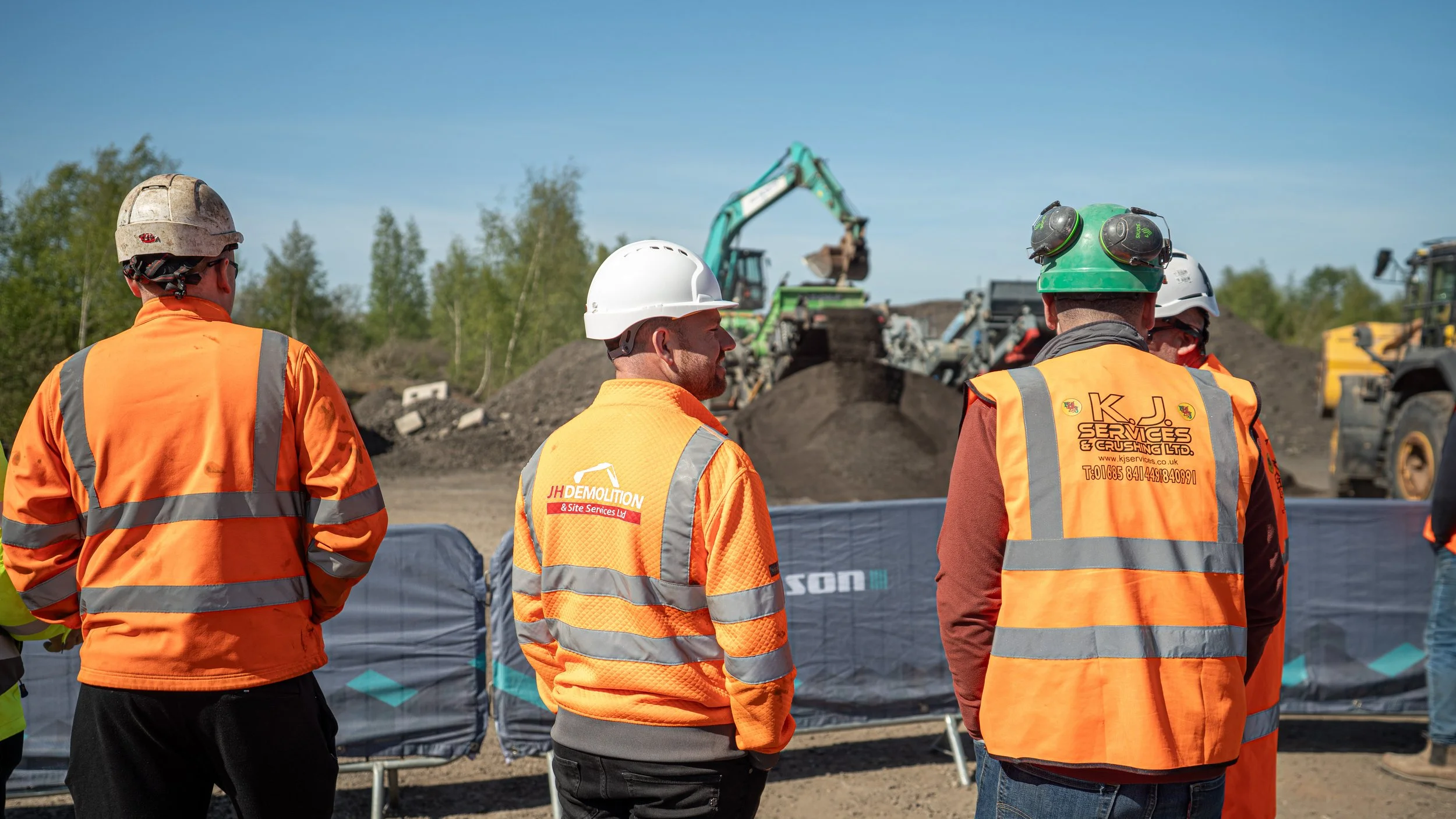 Construction workers wearing orange safety vests and helmets are standing outdoors, observing construction equipment, including excavators, with a background of trees and a blue sky.