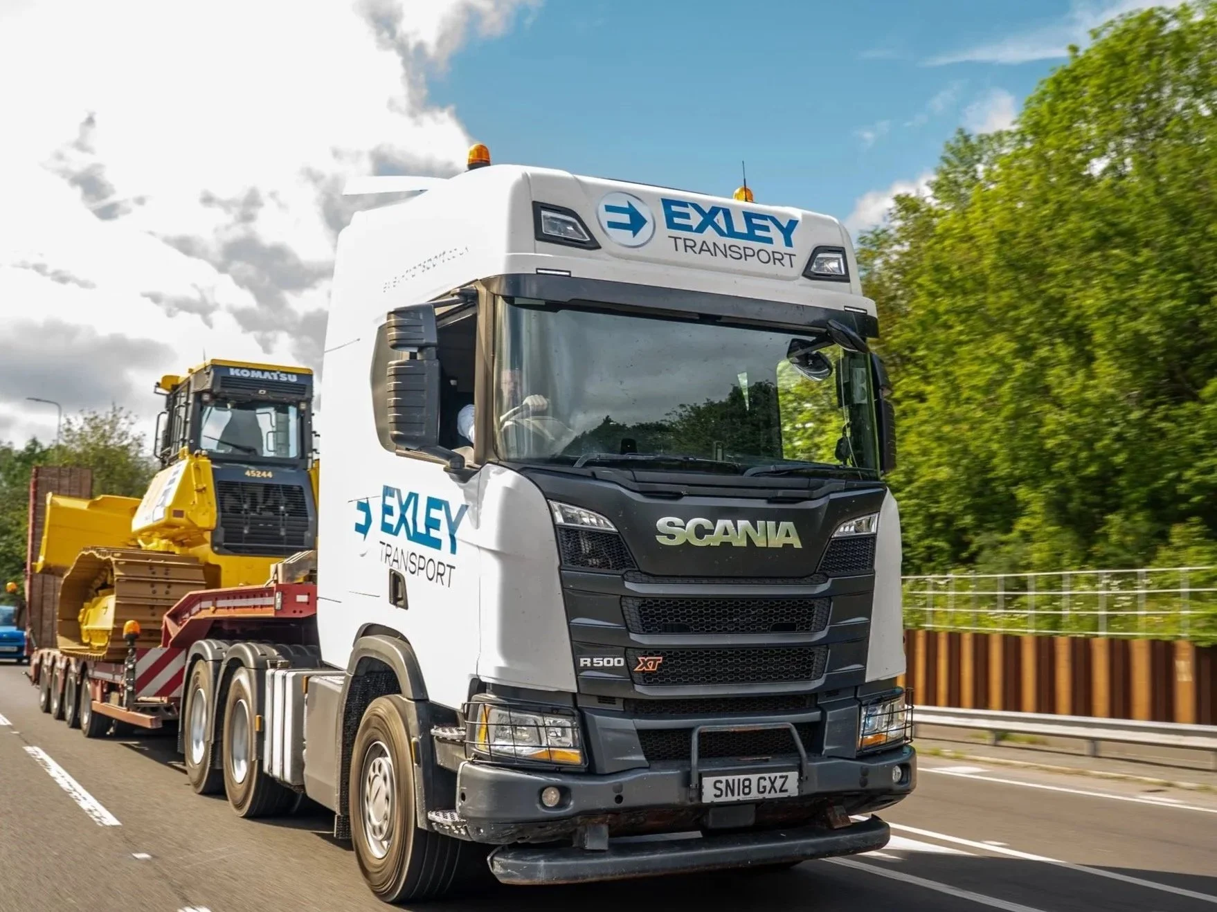 A large truck with a white cab labeled 'EXLEY Transport' on the side is driving on a highway. The truck is pulling a trailer carrying construction equipment, including a yellow excavator. The background shows a partly cloudy sky and green trees.