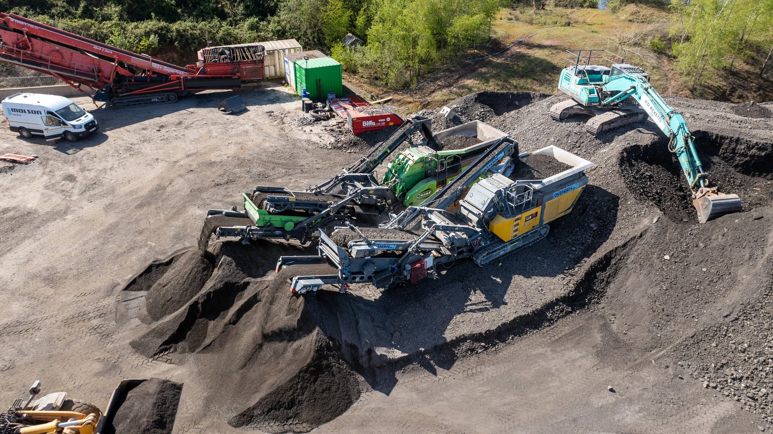 Construction site with heavy machinery, including a large green and black conveyor system, an excavator, and a delivery van, surrounded by dirt and soil.