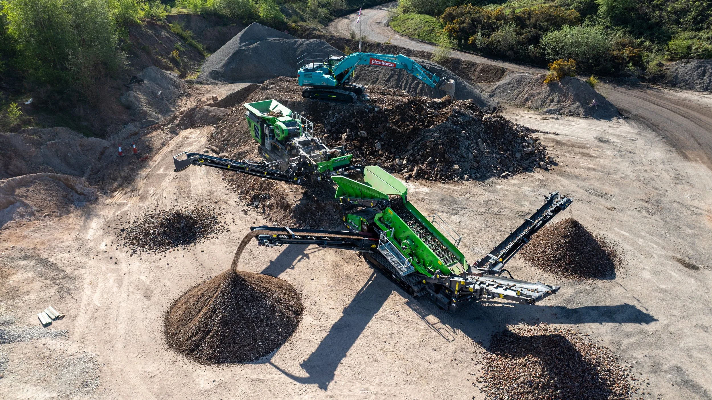 Aerial view of a construction site with heavy machinery, including a green mobile crusher, a blue excavator, and conveyor belts, processing and sorting rocks and gravel on a dirt road surrounded by trees.