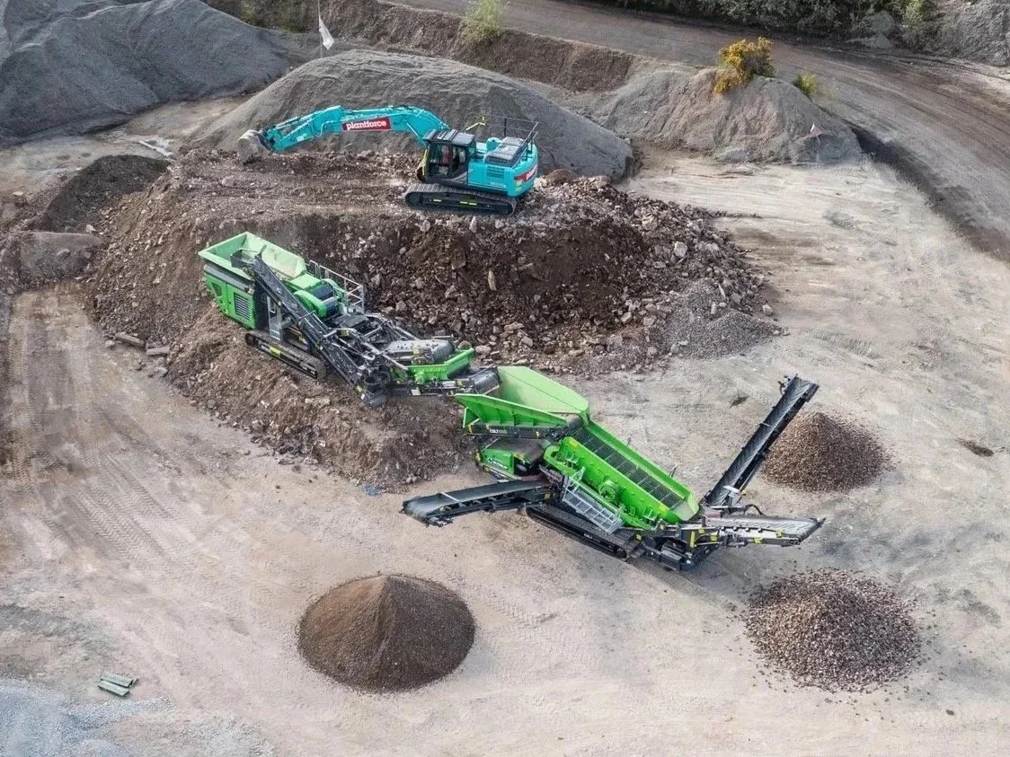 Excavator and mobile crusher machines working on a dirt and gravel site.