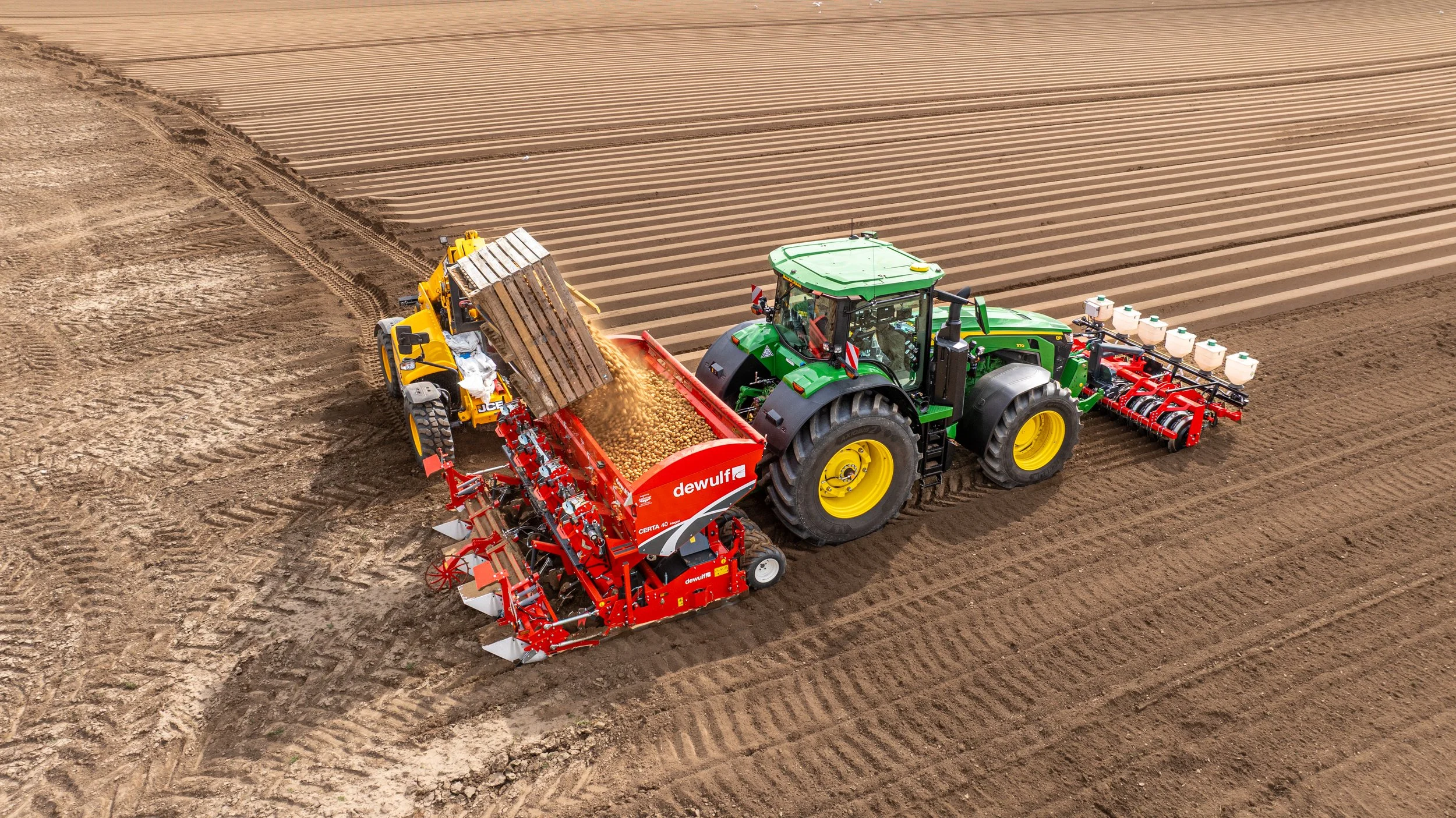 A green tractor and a yellow loader working together on a farm field. The tractor pulls a red seed planter, and the loader deposits seeds onto the soil from a black container. The field has evenly spaced furrows and is prepared for planting.