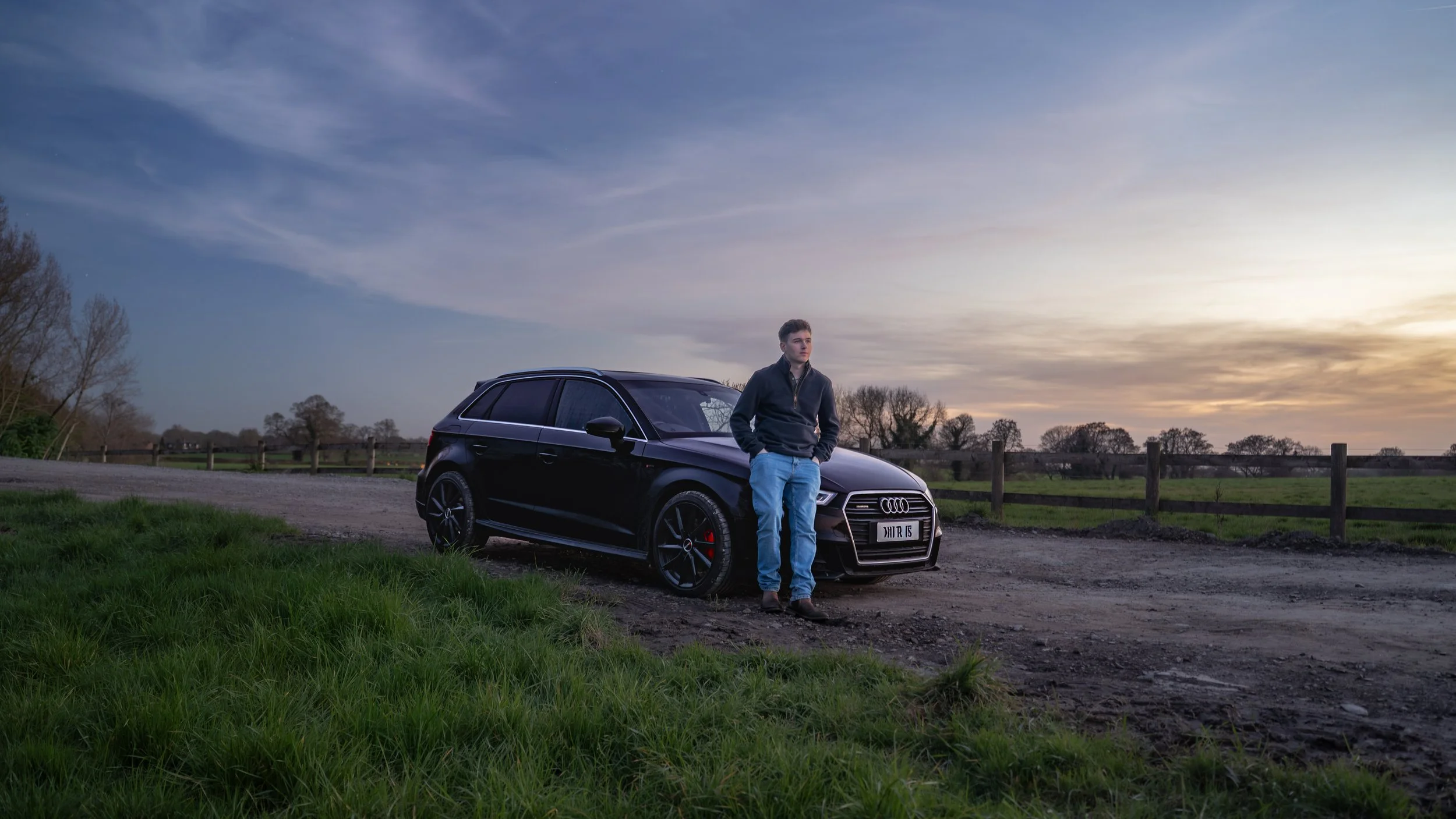 A young man standing next to a black Audi car on a dirt path in a rural area during sunset with a wooden fence and trees in the background.