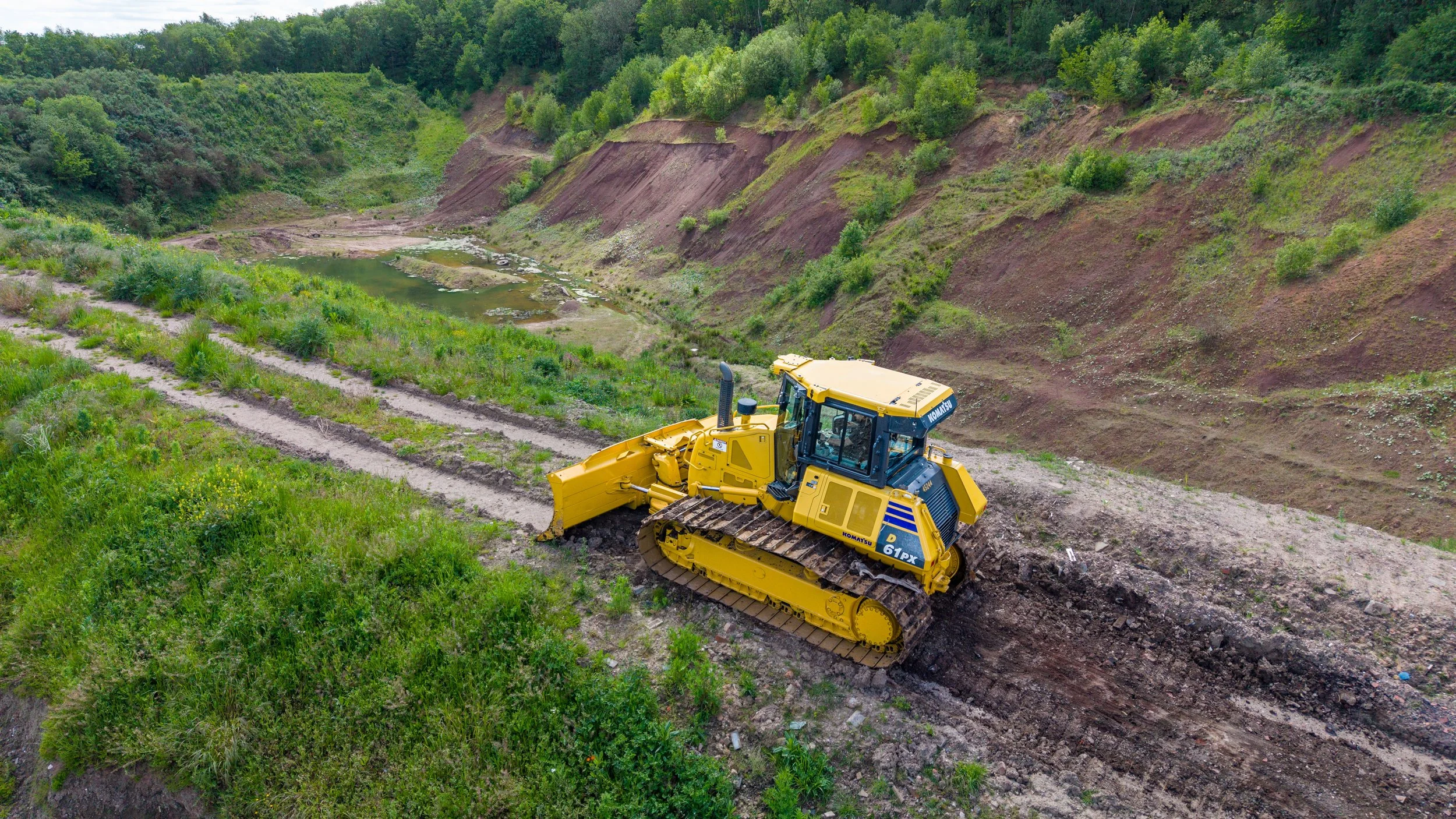 A yellow bulldozer working on a dirt road in a lush green landscape with hills and trees in the background.