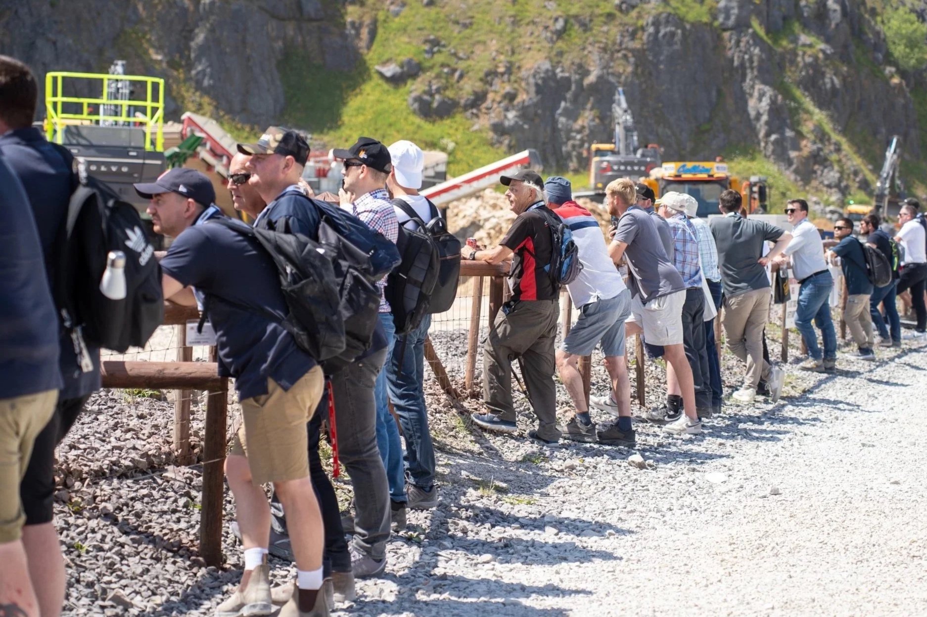 Group of people standing in a line outdoors, leaning on a wooden railing, with construction machinery and rocky hillside in the background.