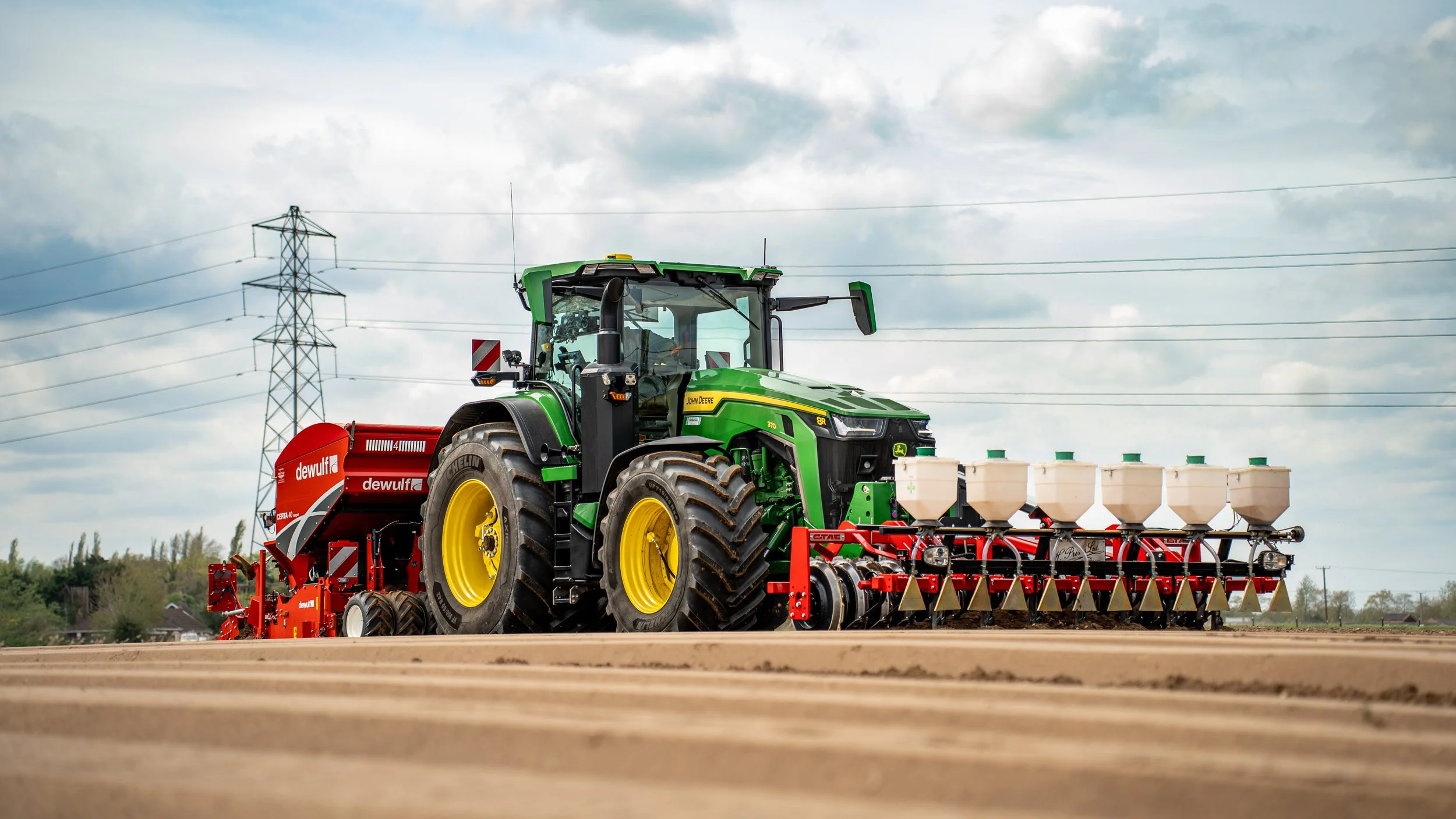 A green John Deere tractor pulling red and white farming equipment in a field under a partly cloudy sky.