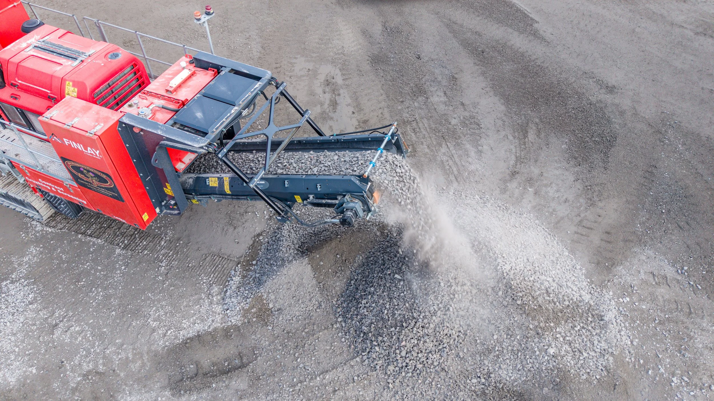 An aerial view of a red industrial machine spreading gravel or dirt on a construction site with a partially prepared dirt surface.