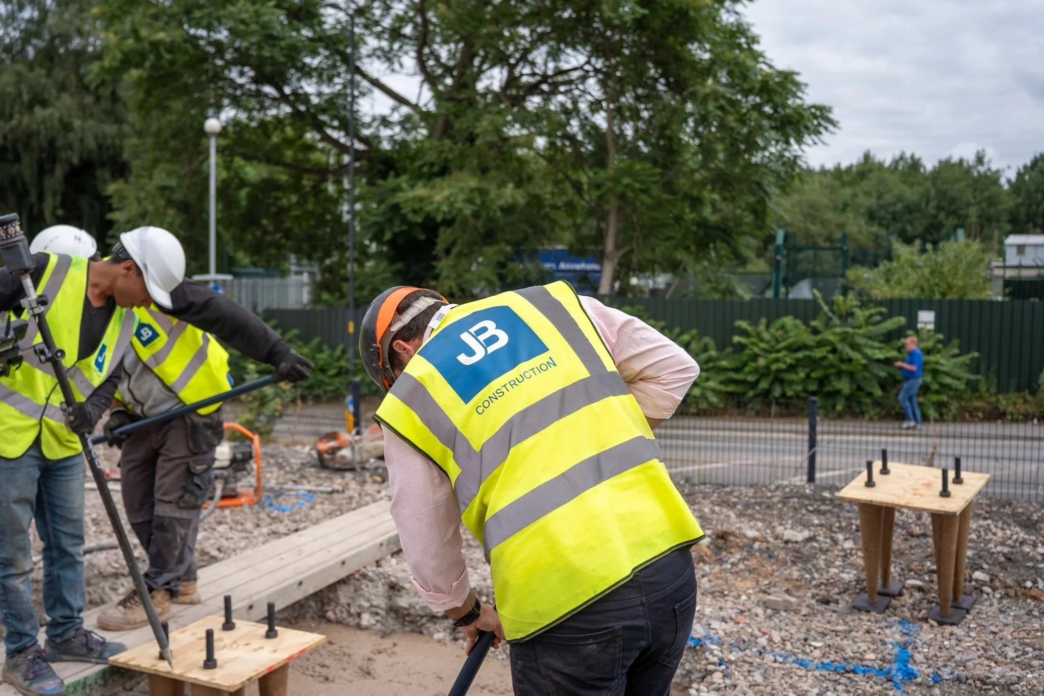 Construction workers in safety vests and helmets working on a site with tools, wooden platforms, and trees in the background.