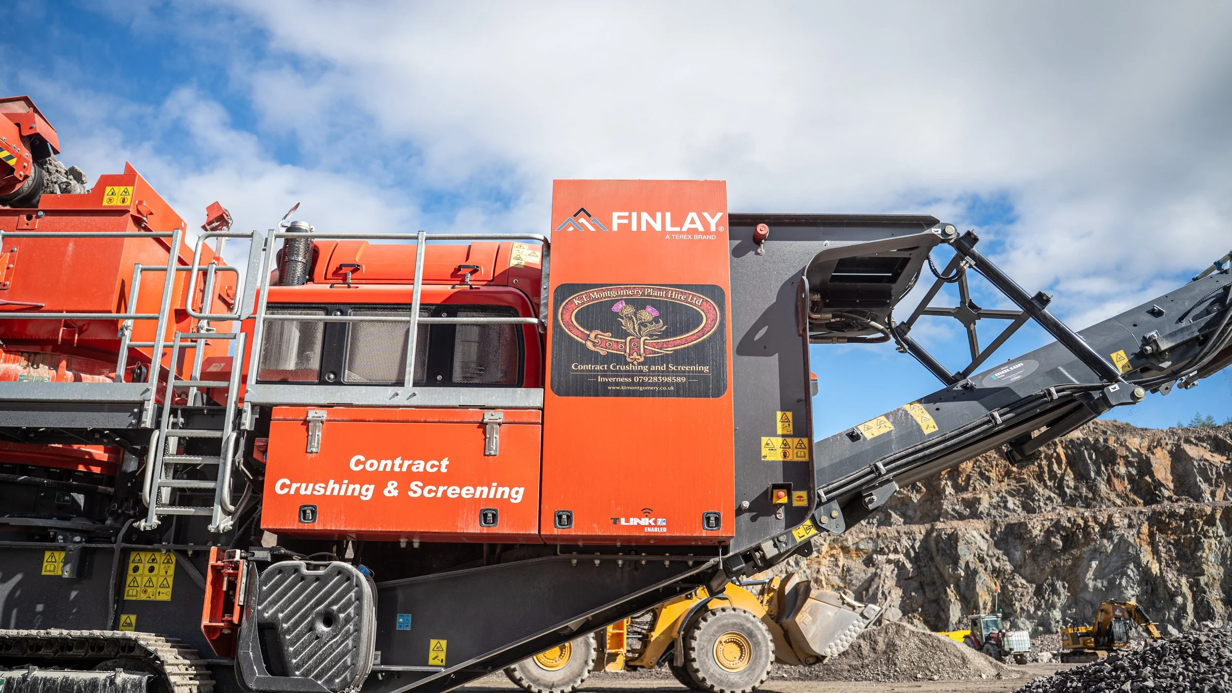 A large orange industrial machine labeled 'Finlay' and 'Contract Crushing & Screening' is operating outdoors with rocky terrain and blue sky with clouds in the background.