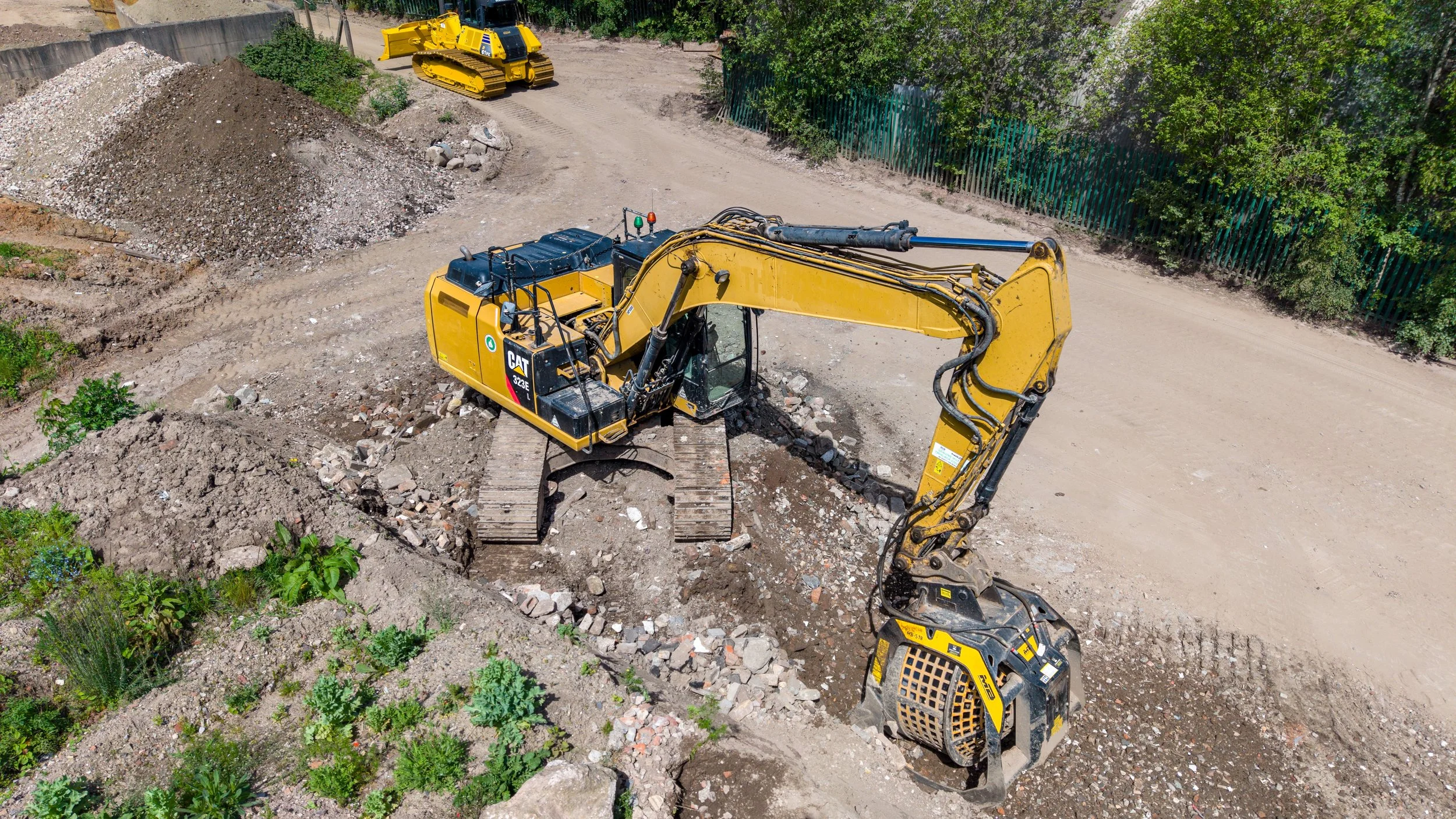 An aerial view of a construction site with a yellow excavator and a yellow bulldozer working on dirt and rocks, with green trees and a fence in the background.