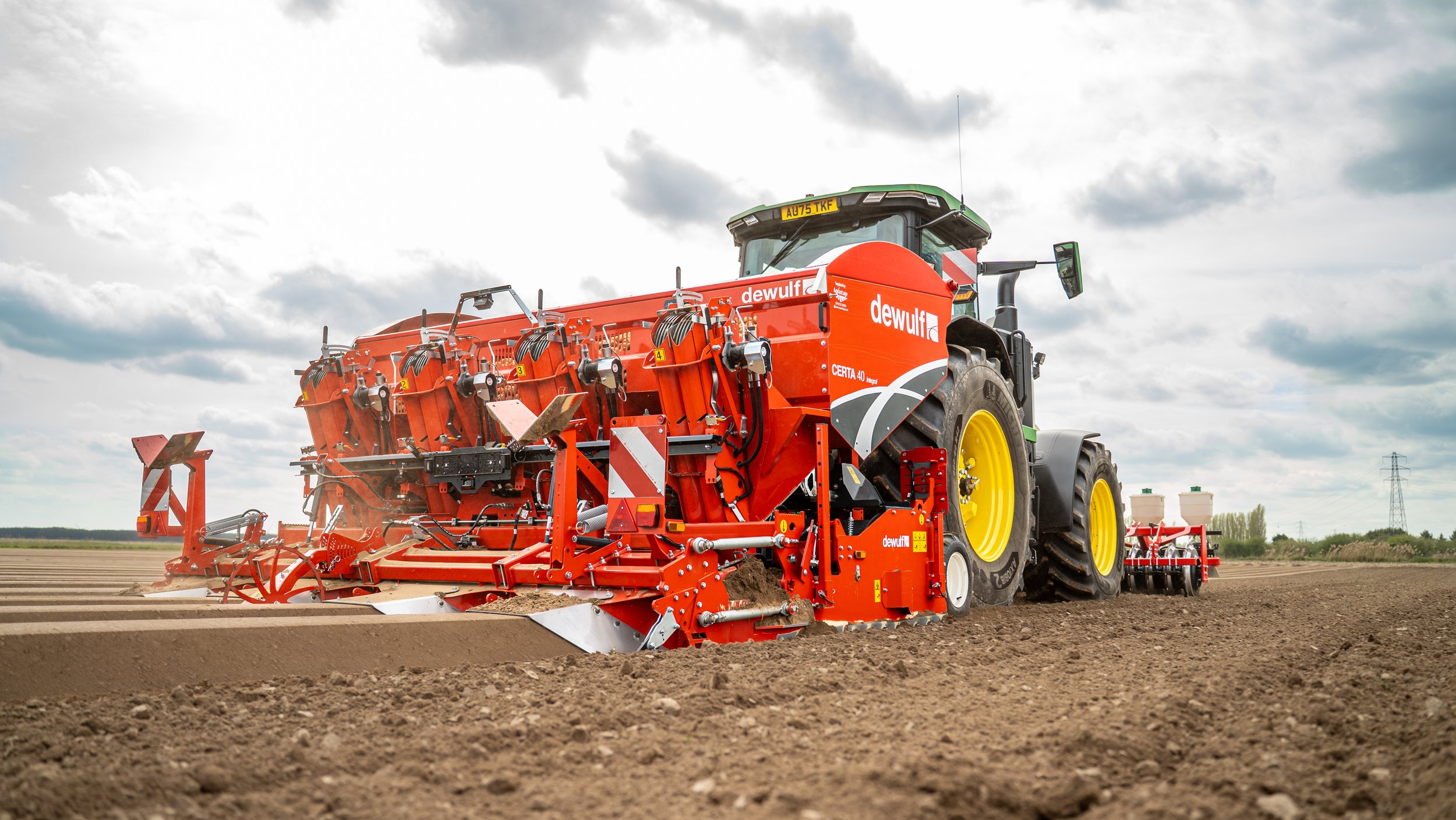 Large green tractor pulling red farming equipment across an open field under a cloudy sky.