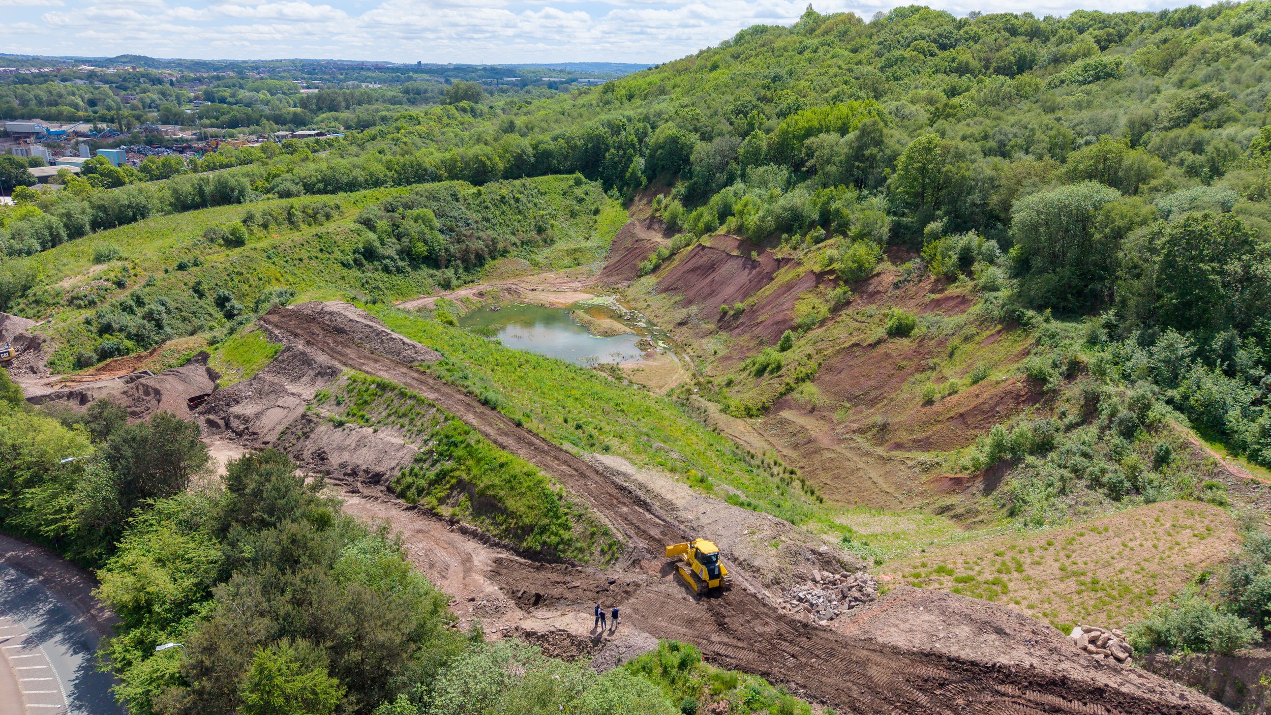 A construction site in a green, hilly area with bulldozers, pathways, and a small pond, blending into a landscape of trees and distant urban buildings under a partly cloudy sky.
