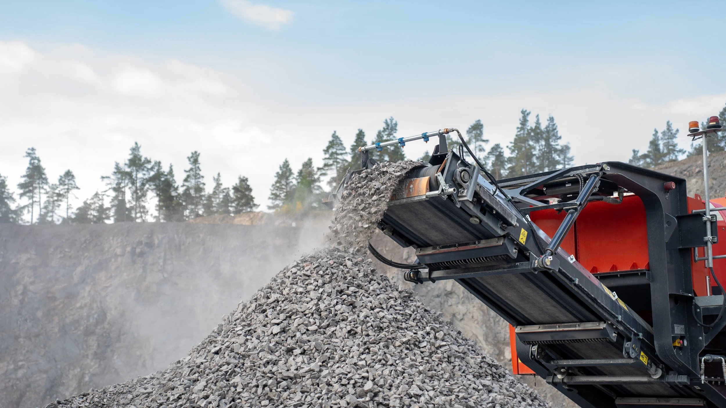 A large industrial crusher machine is crushing rocks into smaller pieces, with a pile of crushed rocks below. The machine is in an outdoor setting with trees and a cloudy sky in the background.