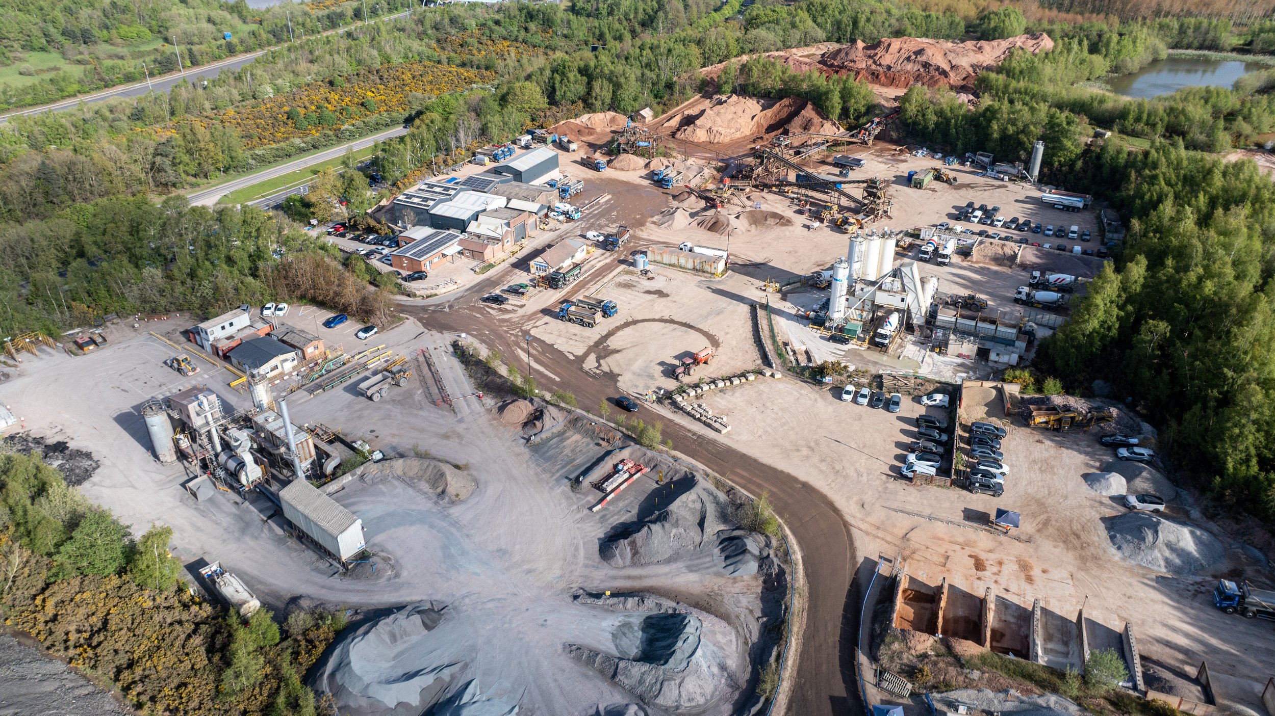 Aerial view of a construction site with machinery, dirt piles, parked vehicles, and surrounded by wooded areas and a road.