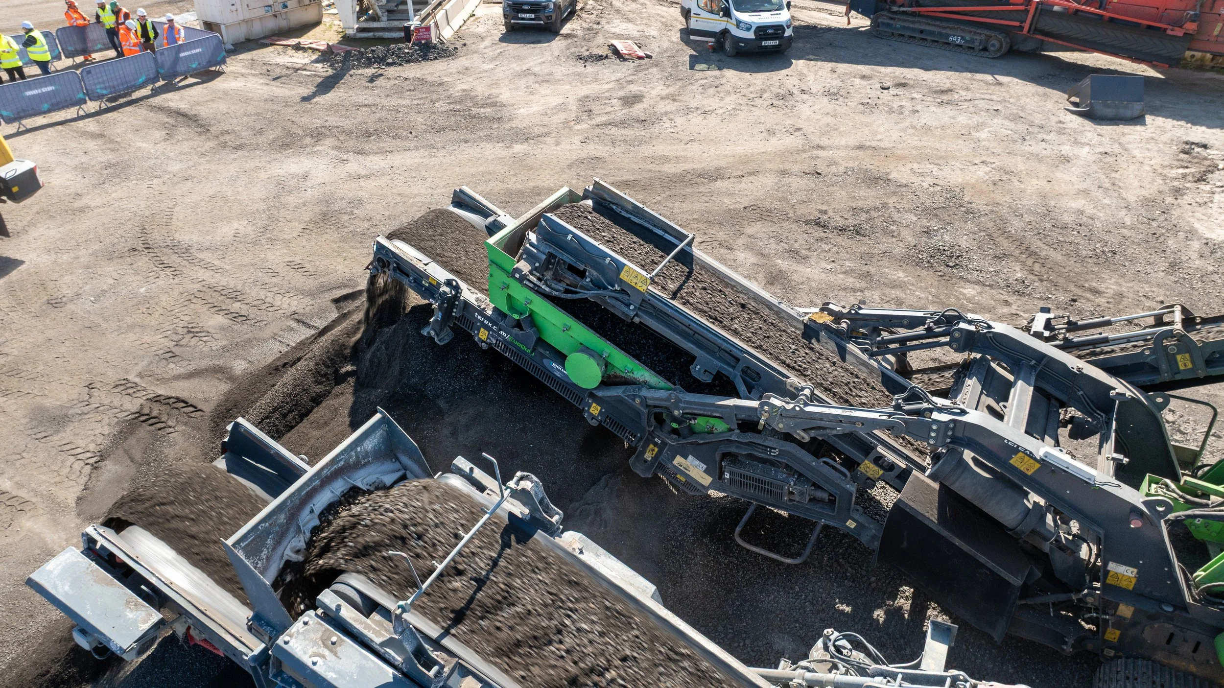 Construction site with multiple workers in safety vests and helmets, and a large green and gray industrial machine moving dirt on a gravel surface.