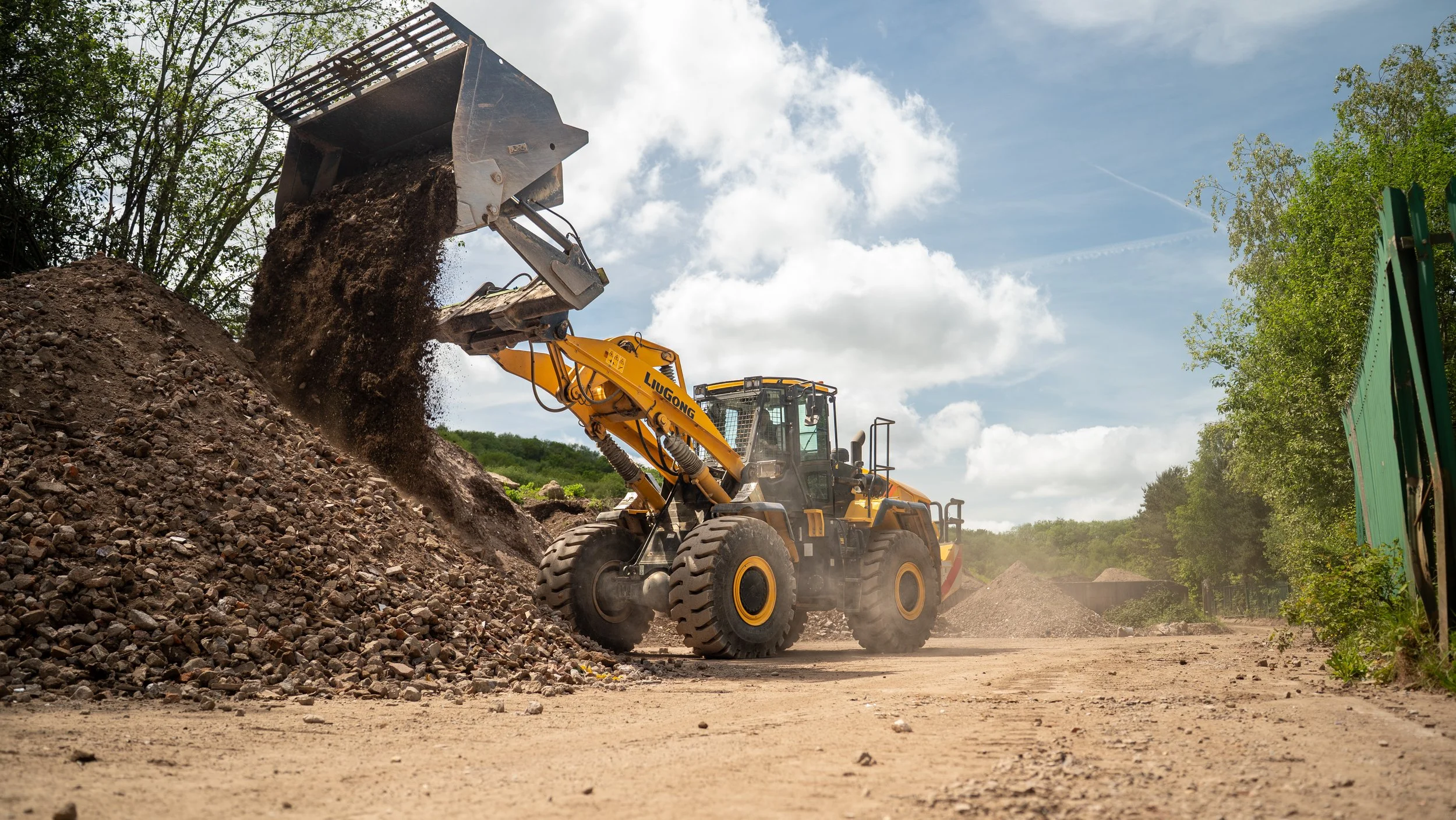 A yellow front loader excavator moving dirt and rocks on a construction site surrounded by trees and blue sky with clouds.