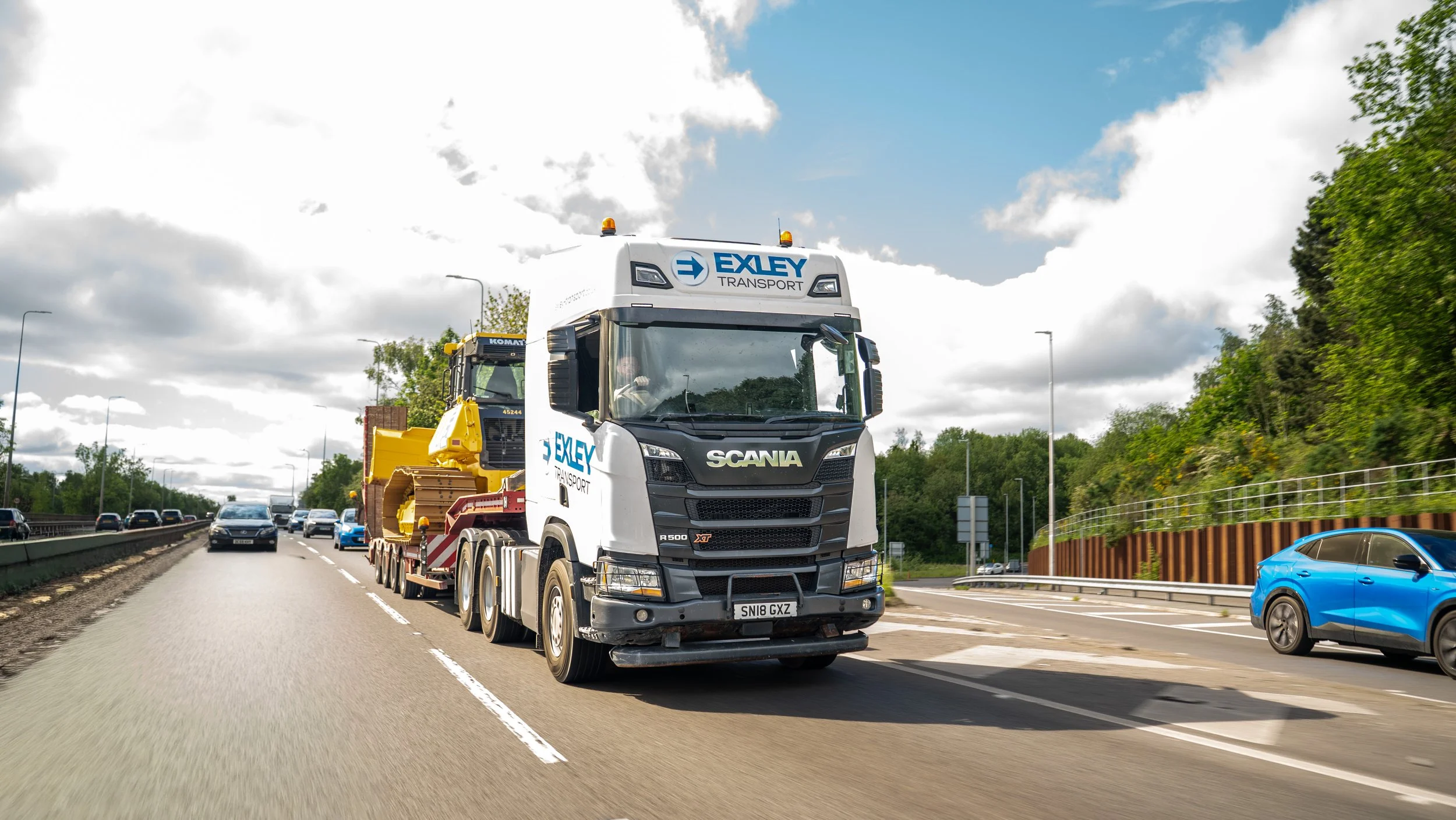 A large white Scania truck from Exley Transport is driving on a highway, carrying construction equipment. Several cars are visible on the road, with trees and a blue sky with clouds in the background.