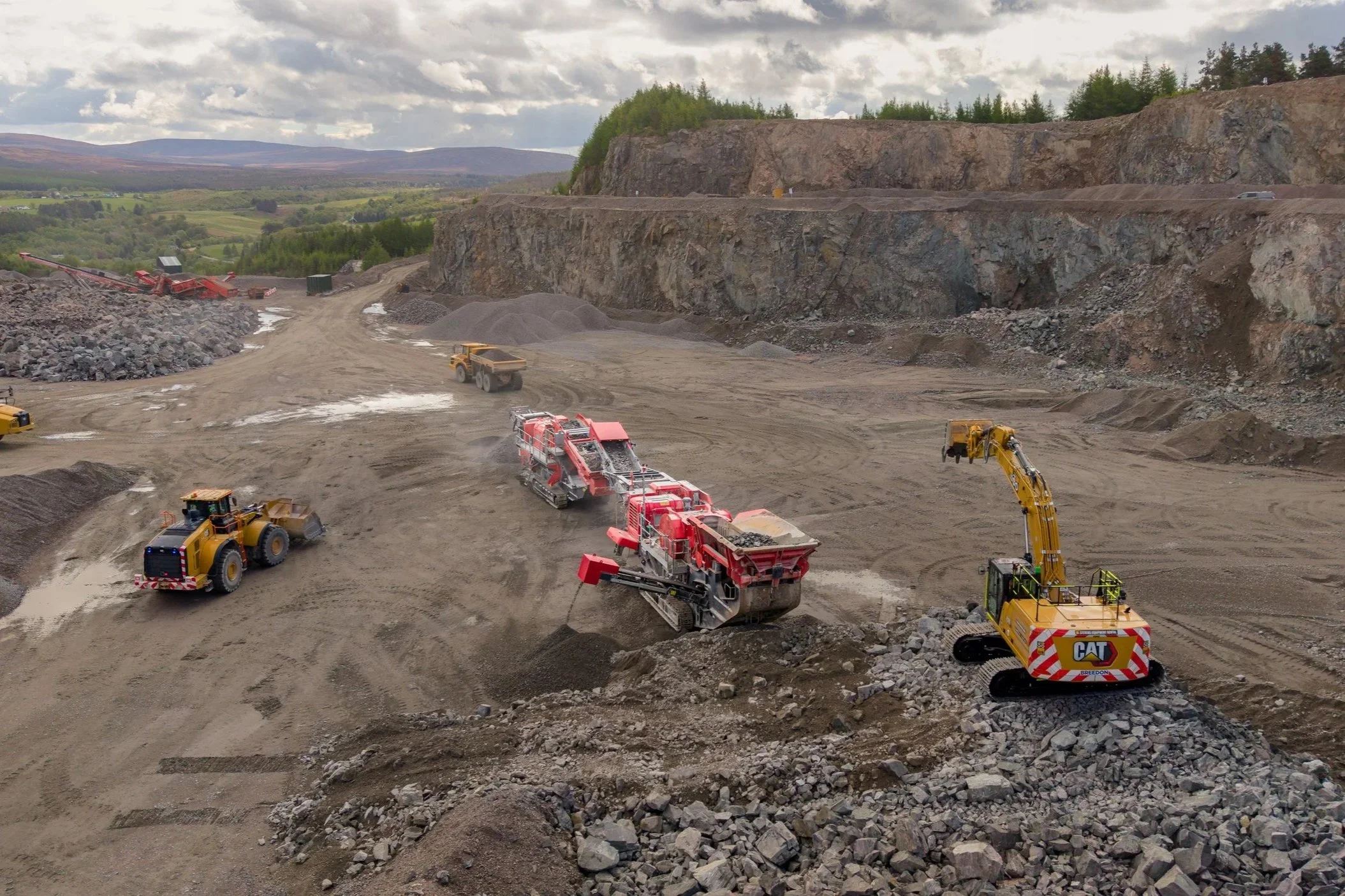 Construction site with heavy machinery working on a rocky terrain, including bulldozers, excavators, and rock crushers, with a backdrop of green hills and cloudy sky.
