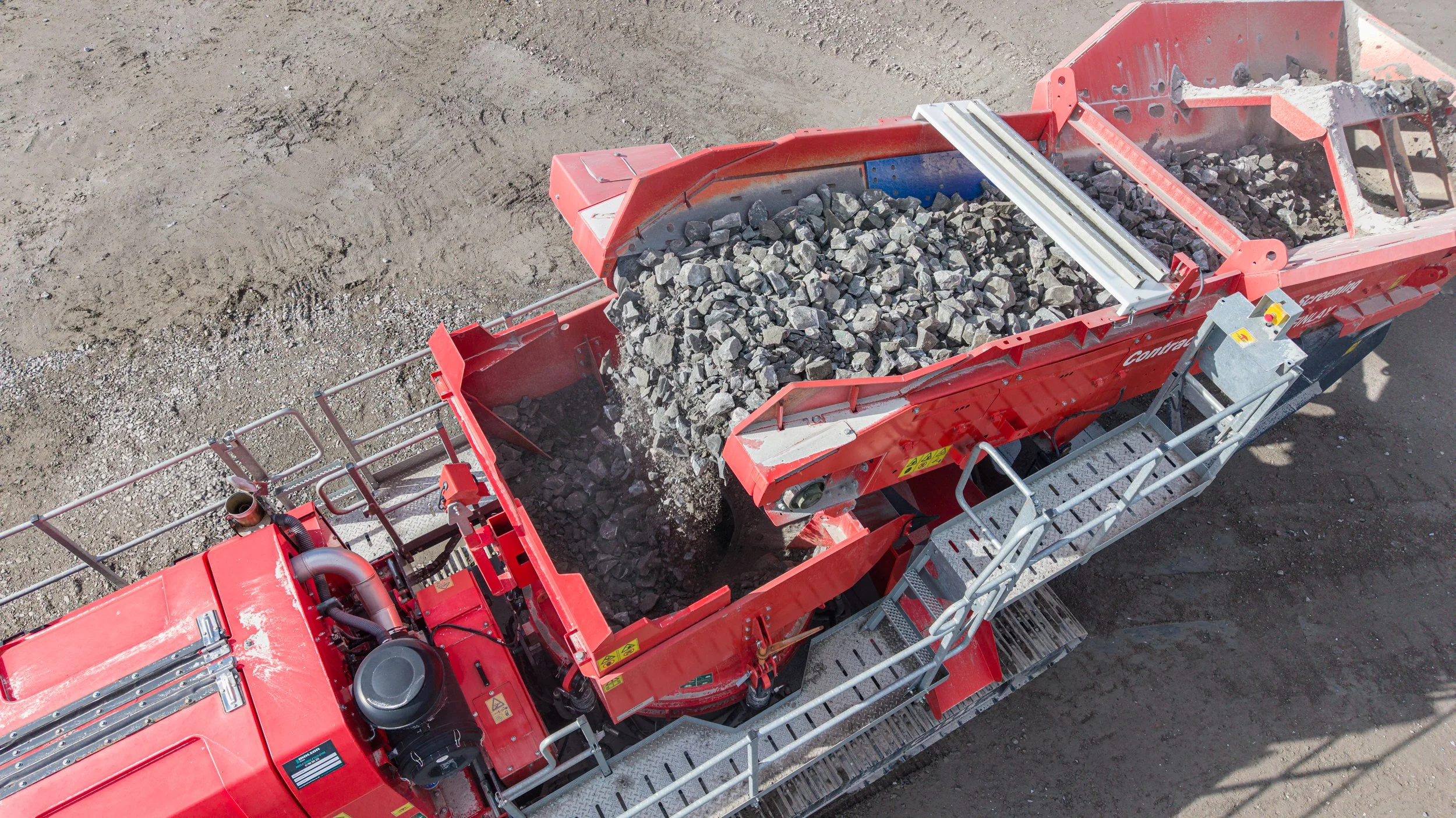 A red industrial machine used for crushing or processing rocks on a construction site, with dirt ground surrounding it.
