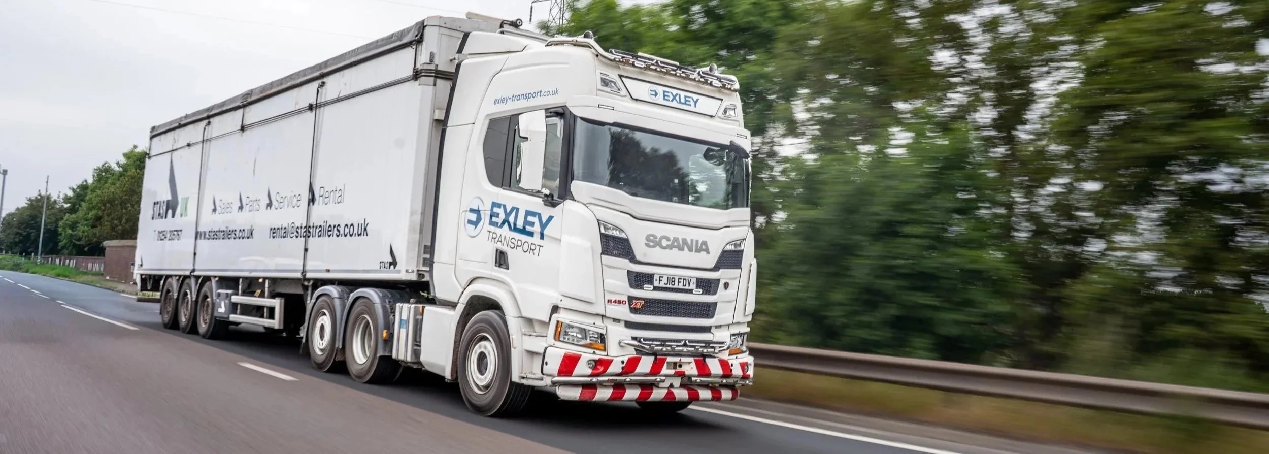 A white semi-truck with the branding 'EXLEY TRANSPORT' driving on a highway next to a guardrail and green trees, with motion blur indicating speed.