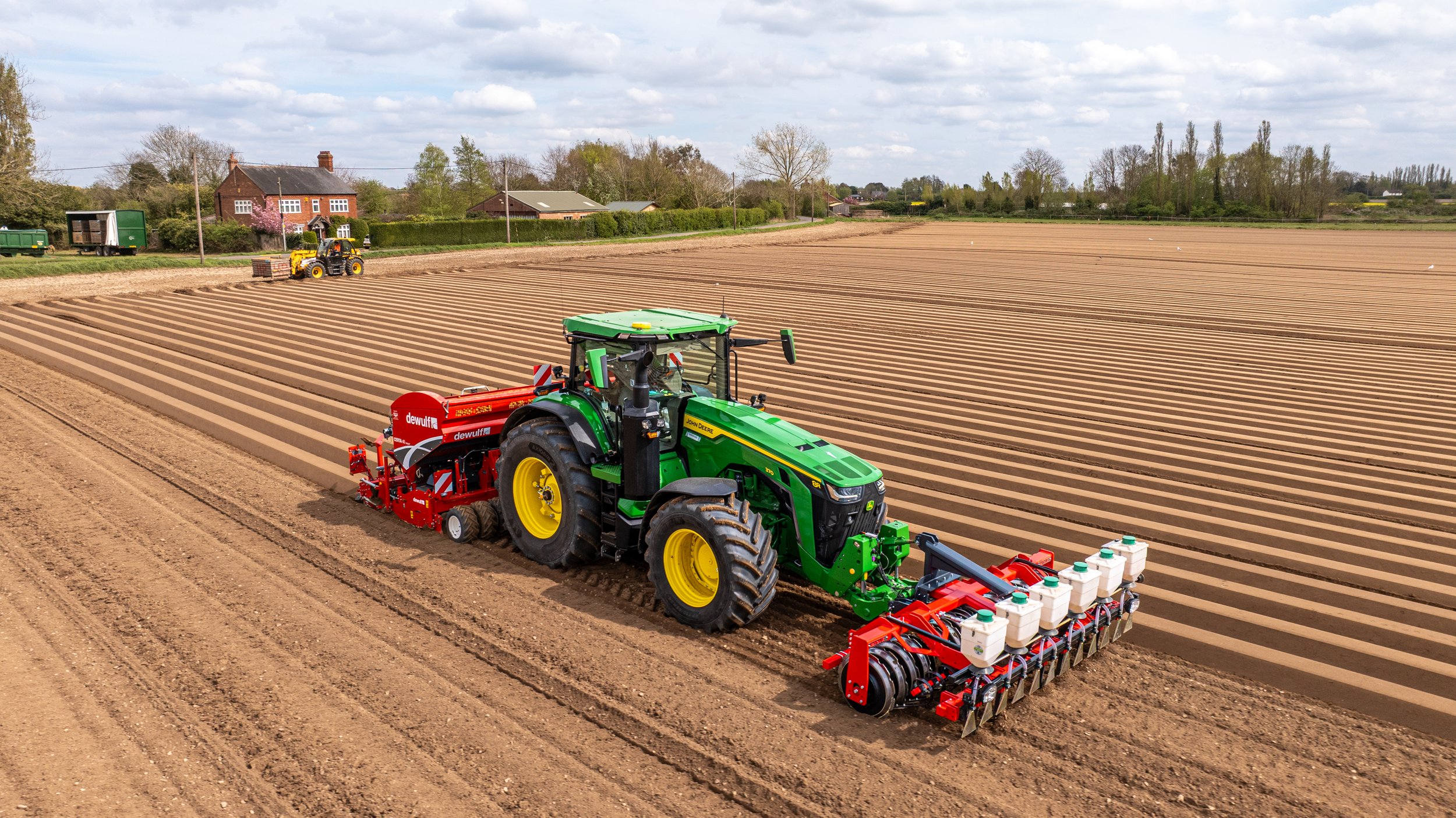 A green tractor working in a large farm field, planting or cultivating rows of soil with a seed drill attached, under a partly cloudy sky.