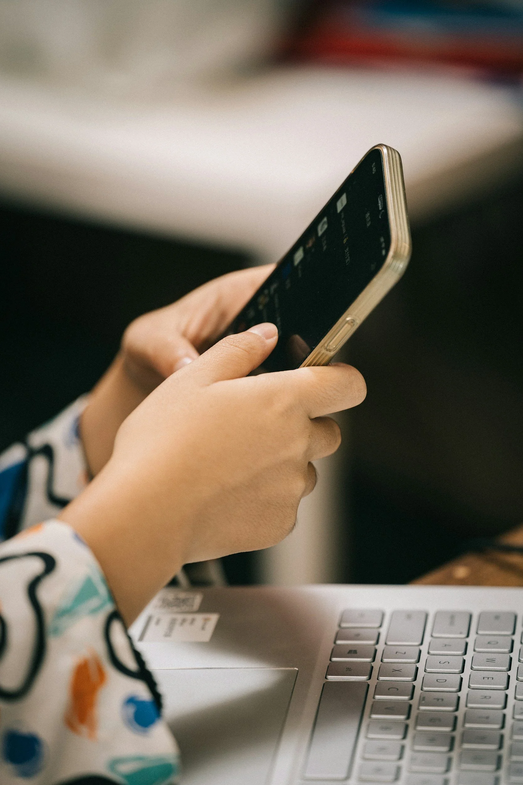 Close-up of a child's hand using a smartphone while sitting at a desk with a silver laptop in the background.